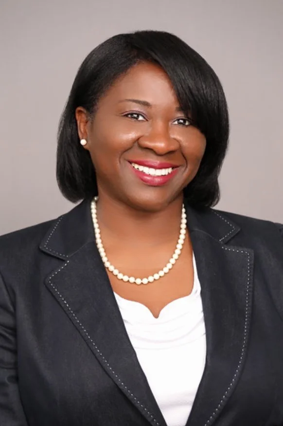 Professional headshot of a smiling Black woman with shoulder-length black hair, wearing a black blazer with white stitching, a white top, pearl earrings, and a pearl necklace against a gray background.