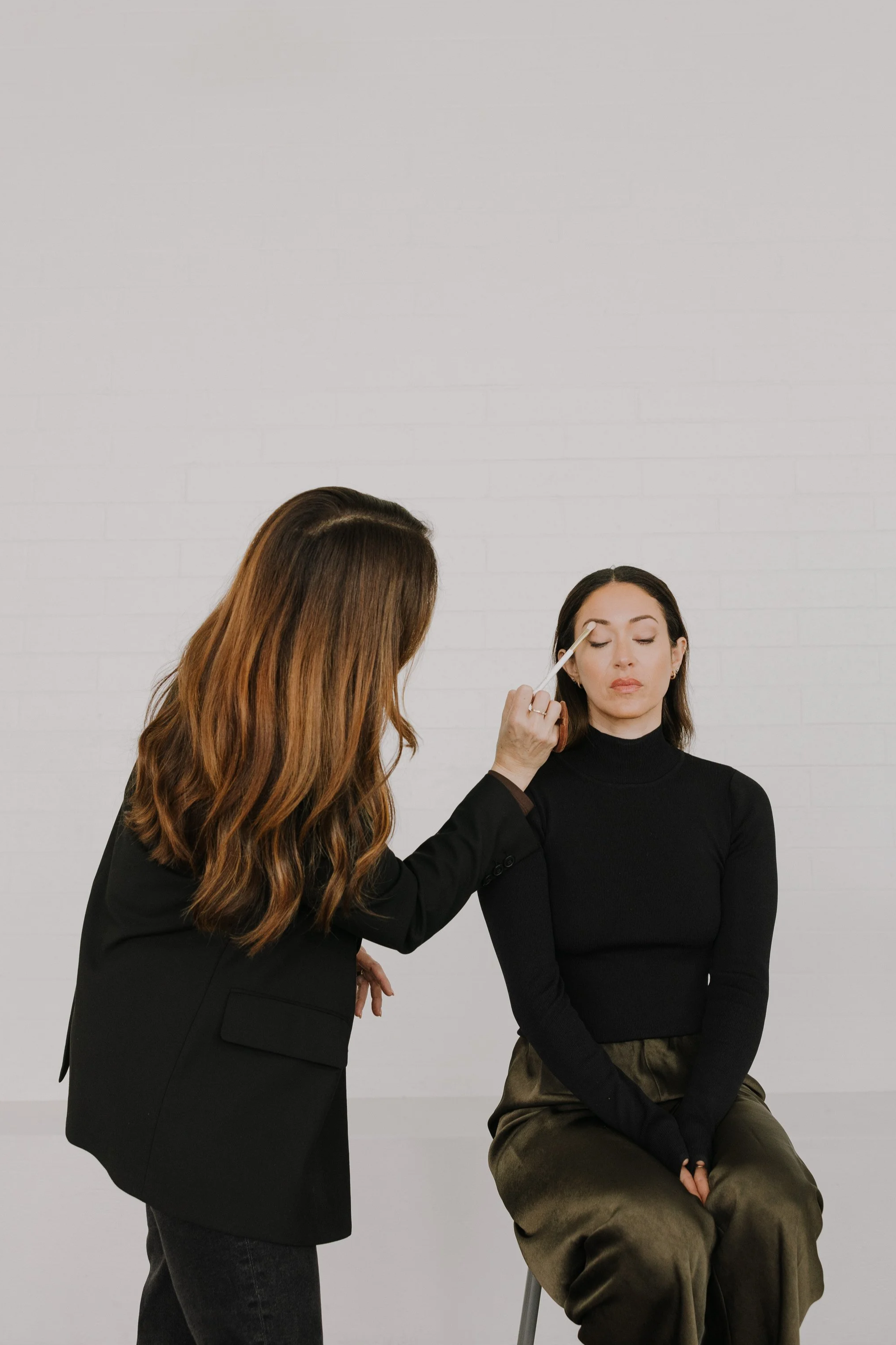 A woman with long brown hair sitting with her eyes closed while another woman with long wavy brown hair applies makeup to her eyelids with a brush.