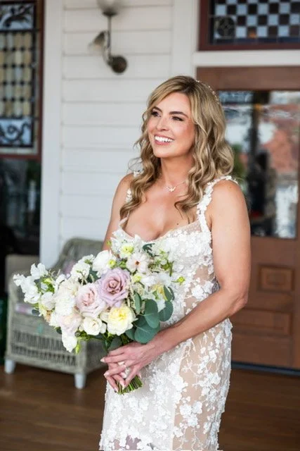 Smiling mature bride with soft Hollywood glam makeup and loose waves, wearing
a lace gown and holding a blush and cream bouquet on the veranda of Hotel del Coronado
in Coronado, CA