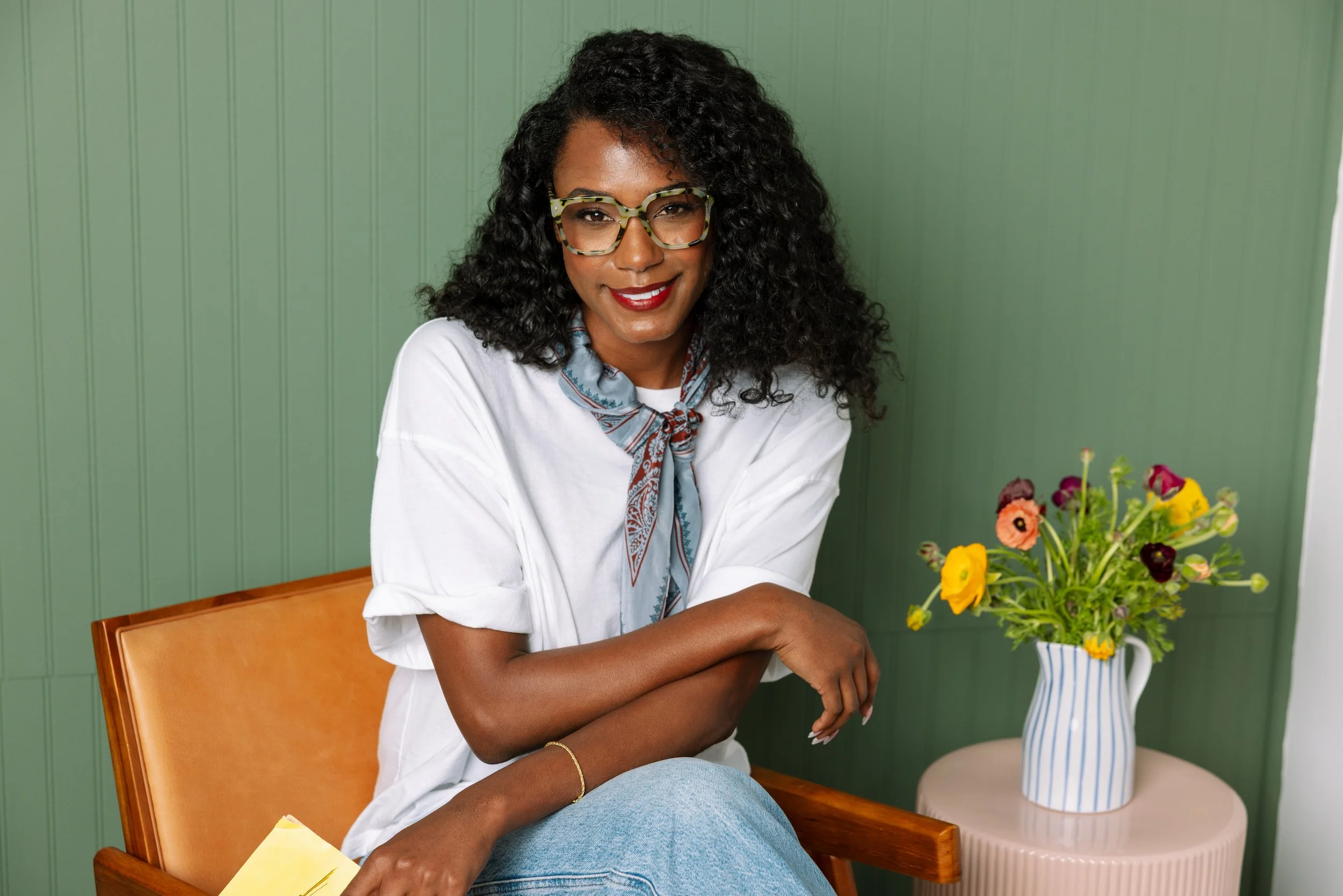 A woman with curly black hair, glasses, and a red lipstick, sitting on an orange chair, smiling, with a green wall in the background, next to a round pink table with a striped ceramic pitcher holding yellow, purple, and pink flowers.