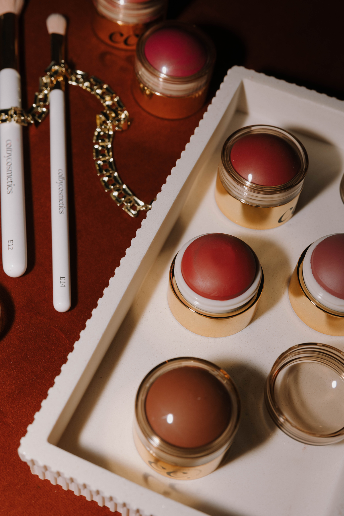 A white palette holds several small containers of lipstick and blush in various shades, with makeup brushes and jewelry including a gold chain and two white-handled makeup brushes nearby.