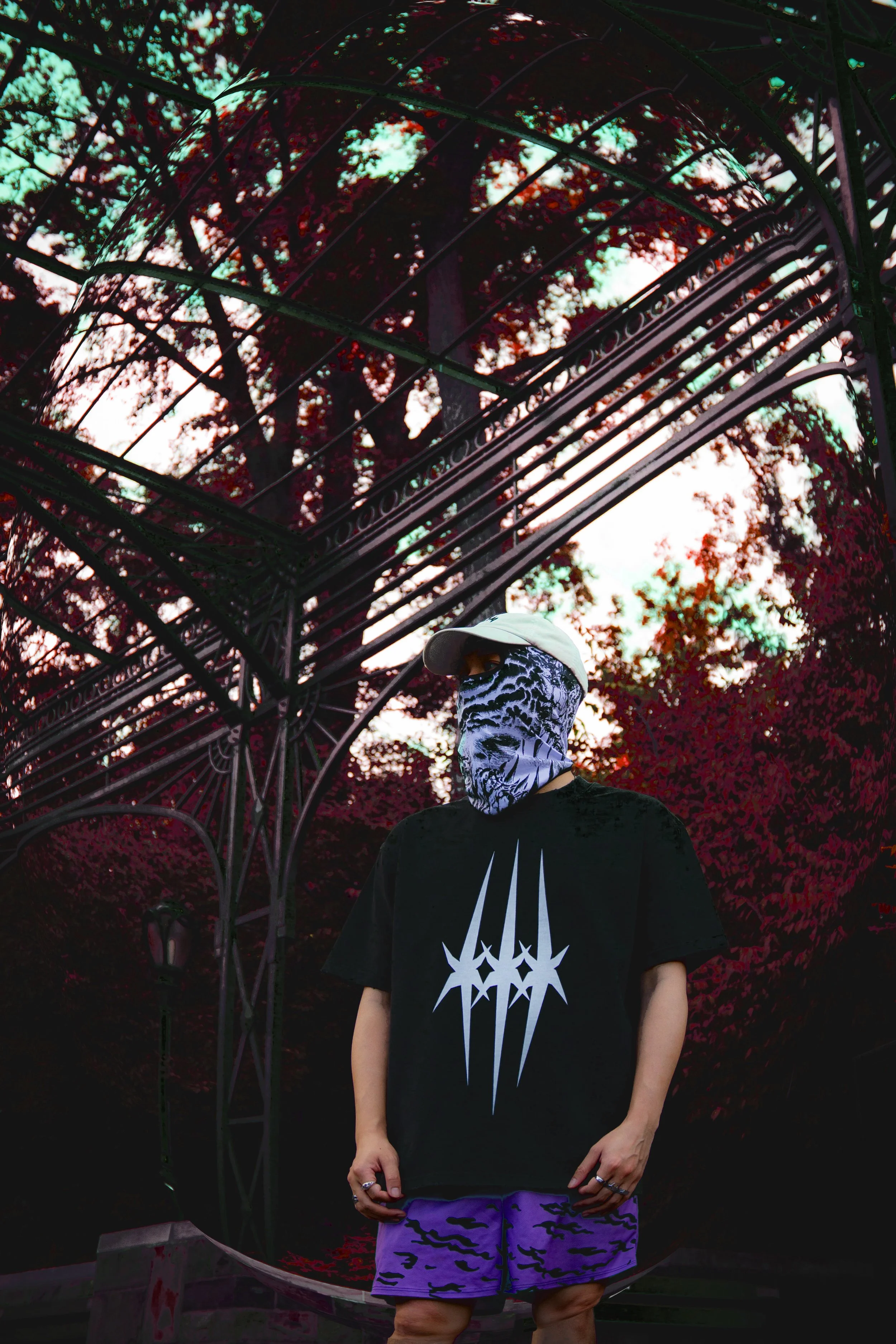 Person wearing a bandana over their face, a baseball cap, a black T-shirt with a white star-like symbol, and purple camouflage shorts standing outdoors under a metal archway with trees and sky in the background.