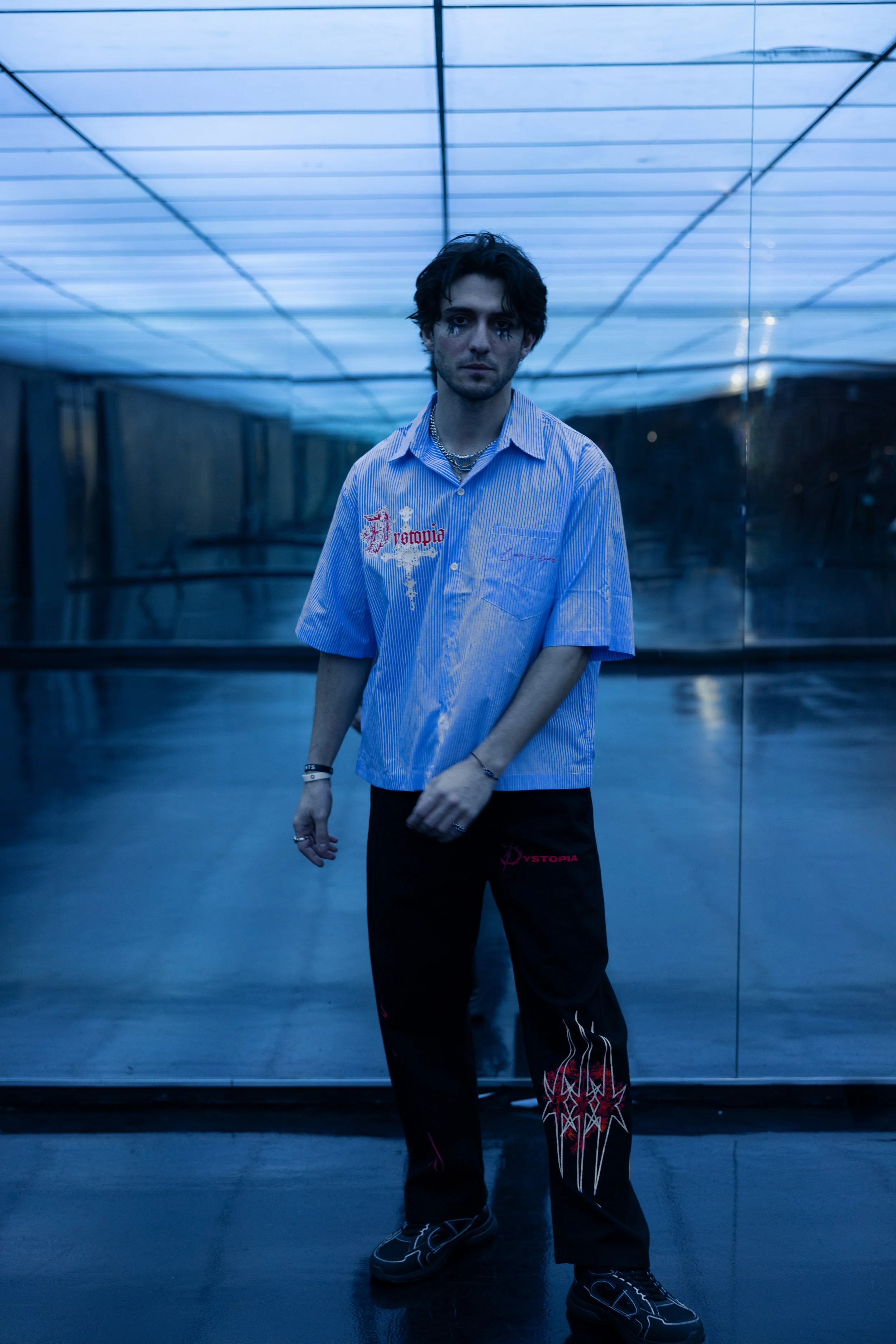 A young man standing in front of a reflective wall with a grid ceiling illuminated with blue lights, wearing a blue striped shirt and black pants with red and white graphic designs.