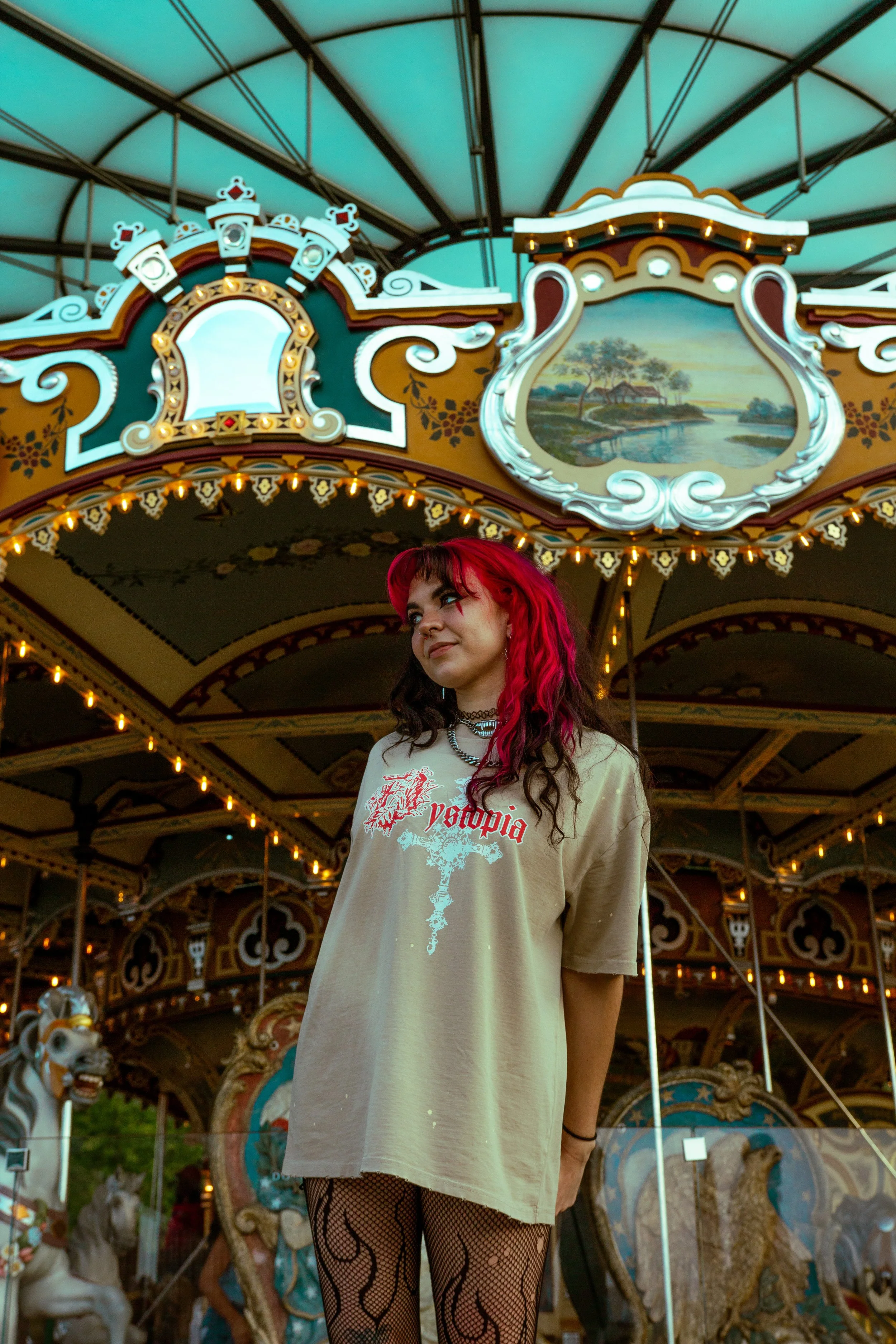 A young woman with red and black hair standing in front of a carousel at an amusement park.