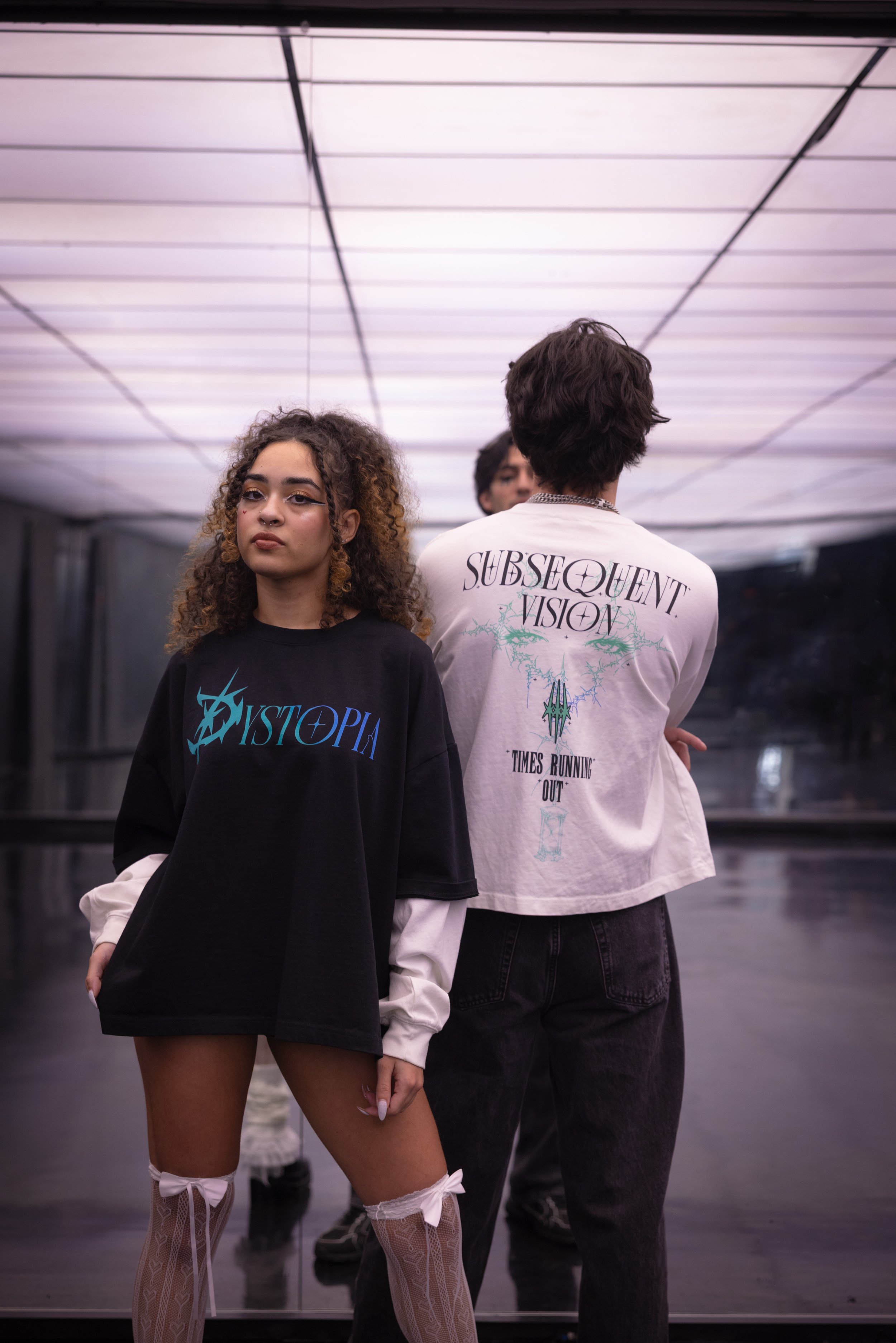 Two young people standing in front of a mirror in a modern room. The woman on the left has curly hair, wears a black oversized t-shirt with blue text, sheer thigh-high stockings with bows. The man on the right has short hair, wears a white oversized 