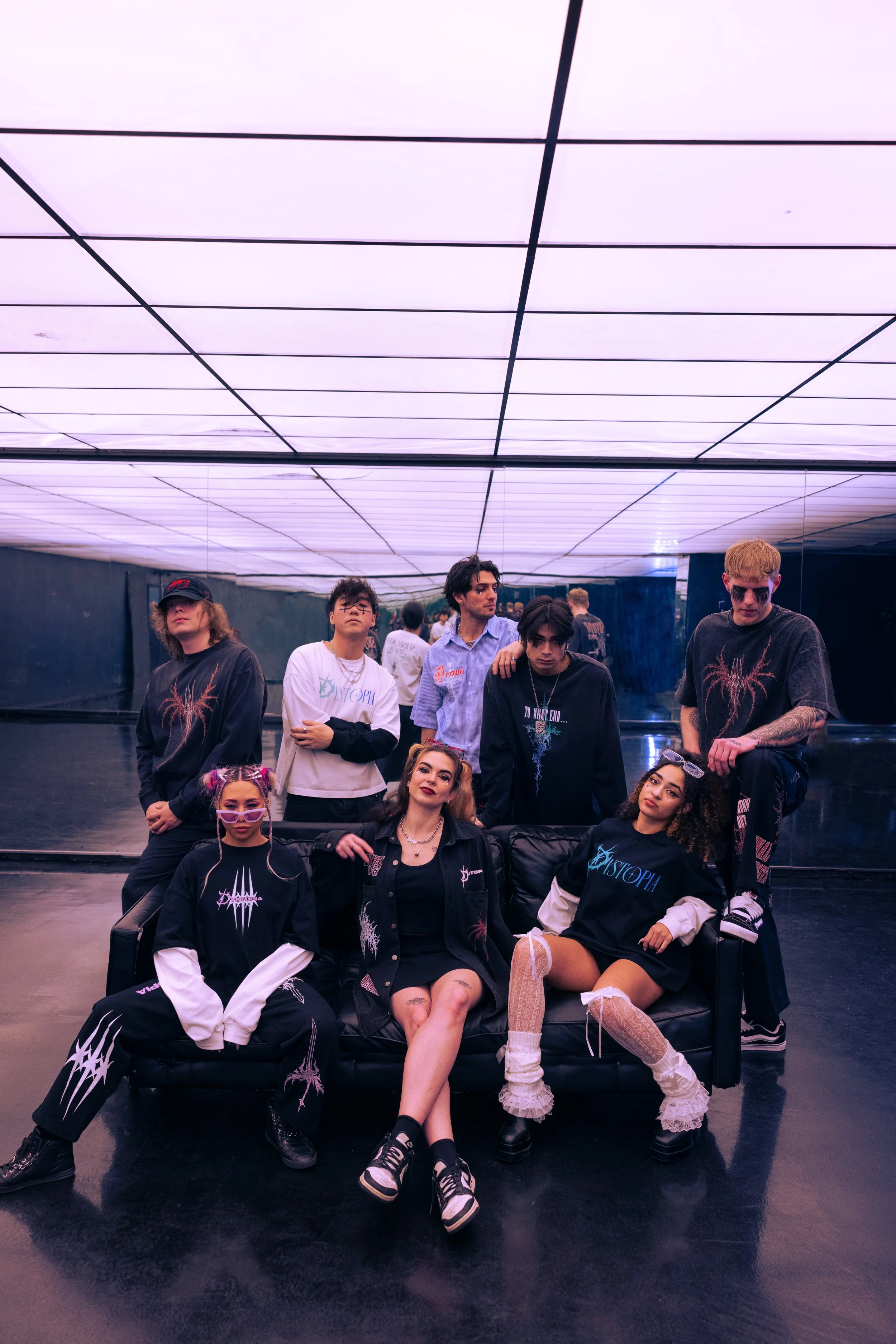 Group of nine young people in dark, edgy clothing with graphic designs, posing in a futuristic, neon-lit room with a reflective floor and ceiling panels.