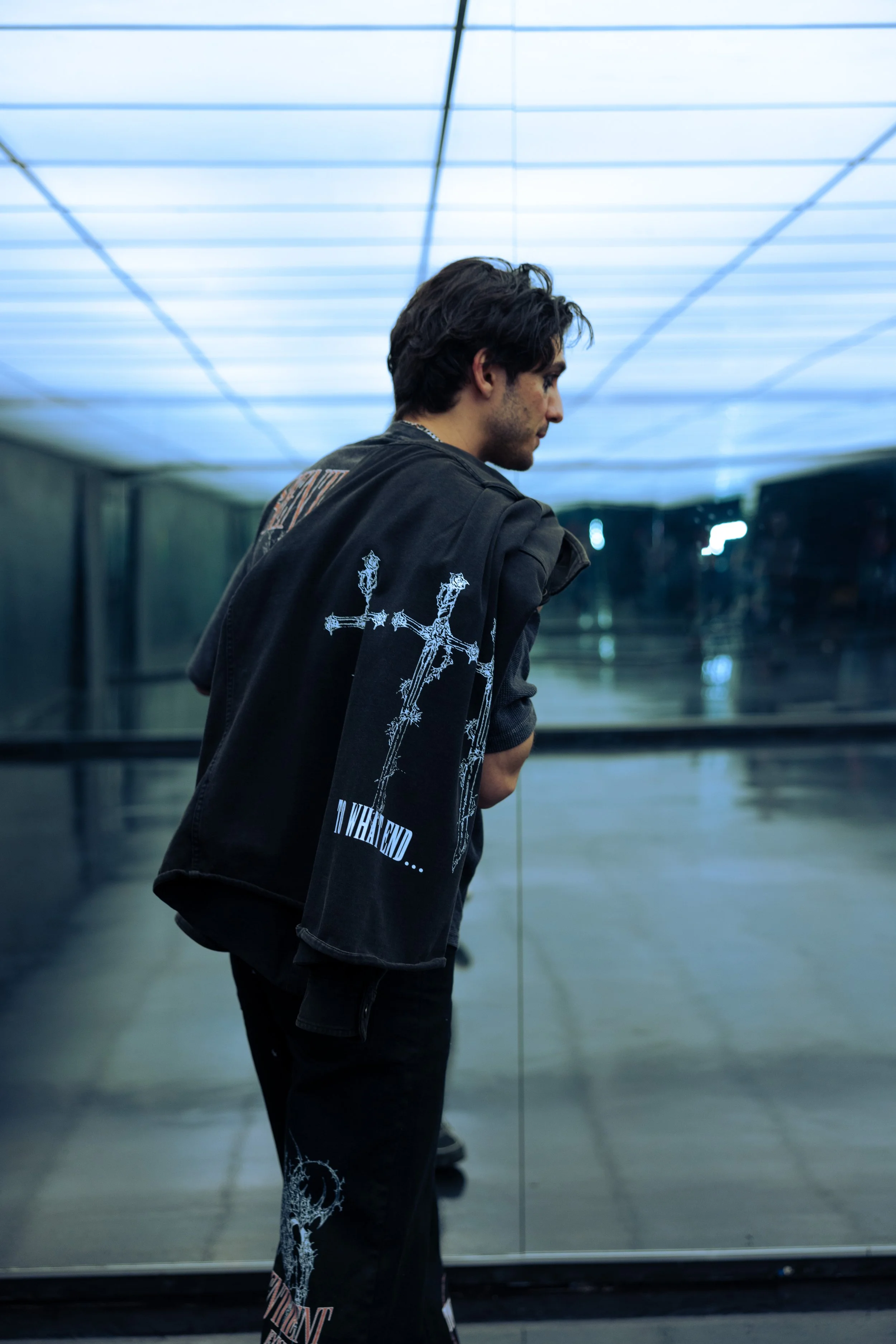 A man standing in front of a mirror with blue lighting ceiling, wearing a black jacket with gothic print and text, with dark hair, looking down, in an indoor modern space.
