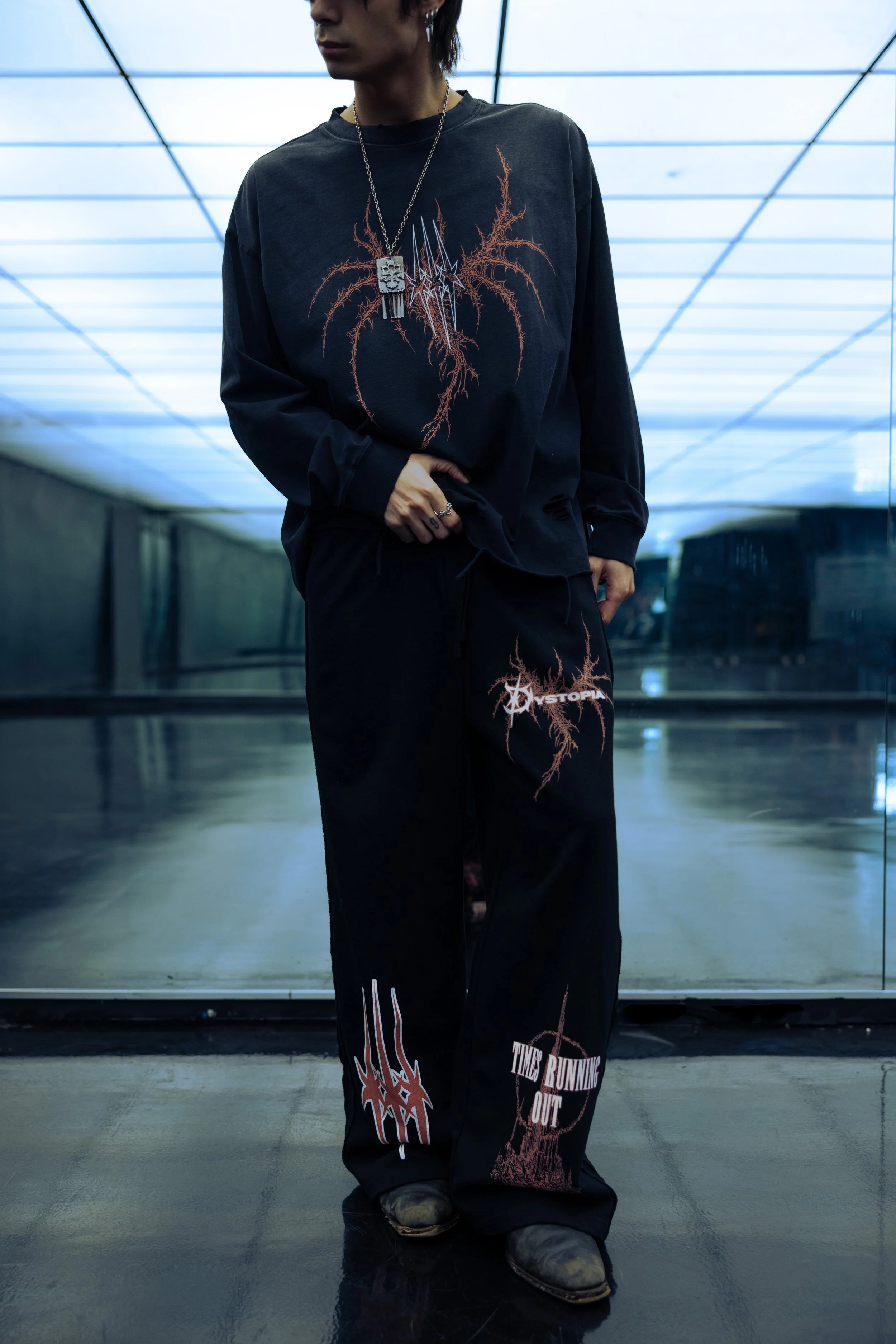 Person wearing black graphic clothing with text and designs, standing in front of a reflective surface, in an underground or modern indoor space.