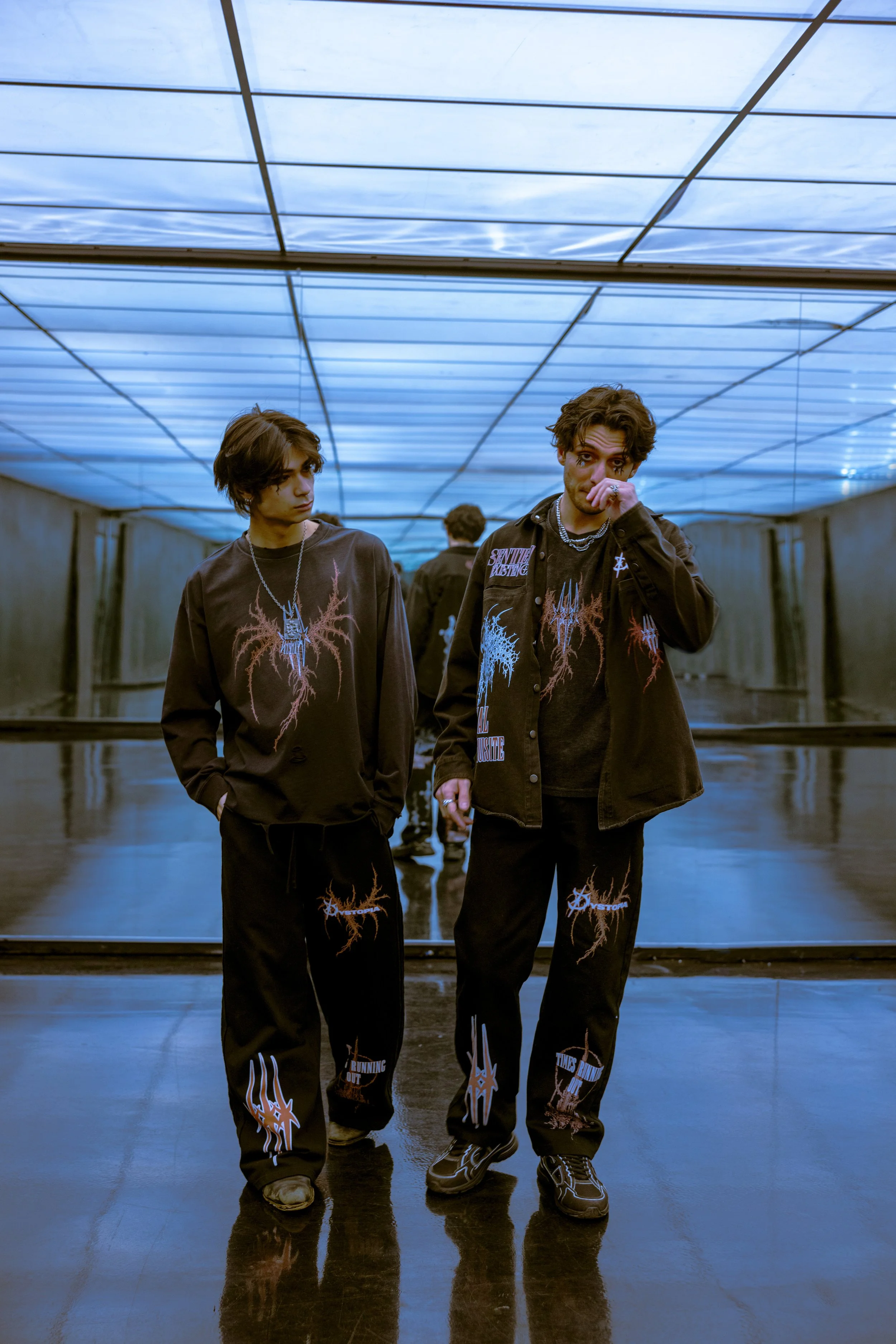 Two young men standing in front of a mirror in a modern, illuminated underground space wearing matching black graphic clothing with abstract designs.