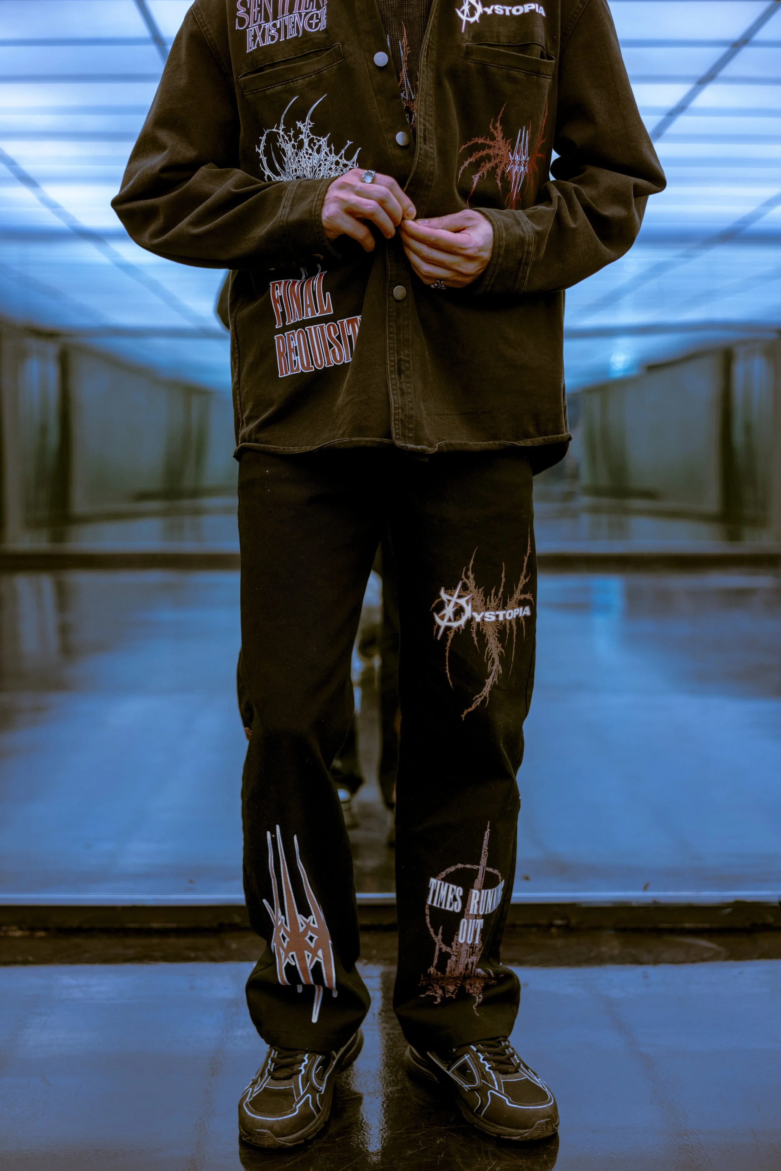 Person wearing black outfit with graphic patches, standing indoors on a reflective floor, with artificial ceiling lights.