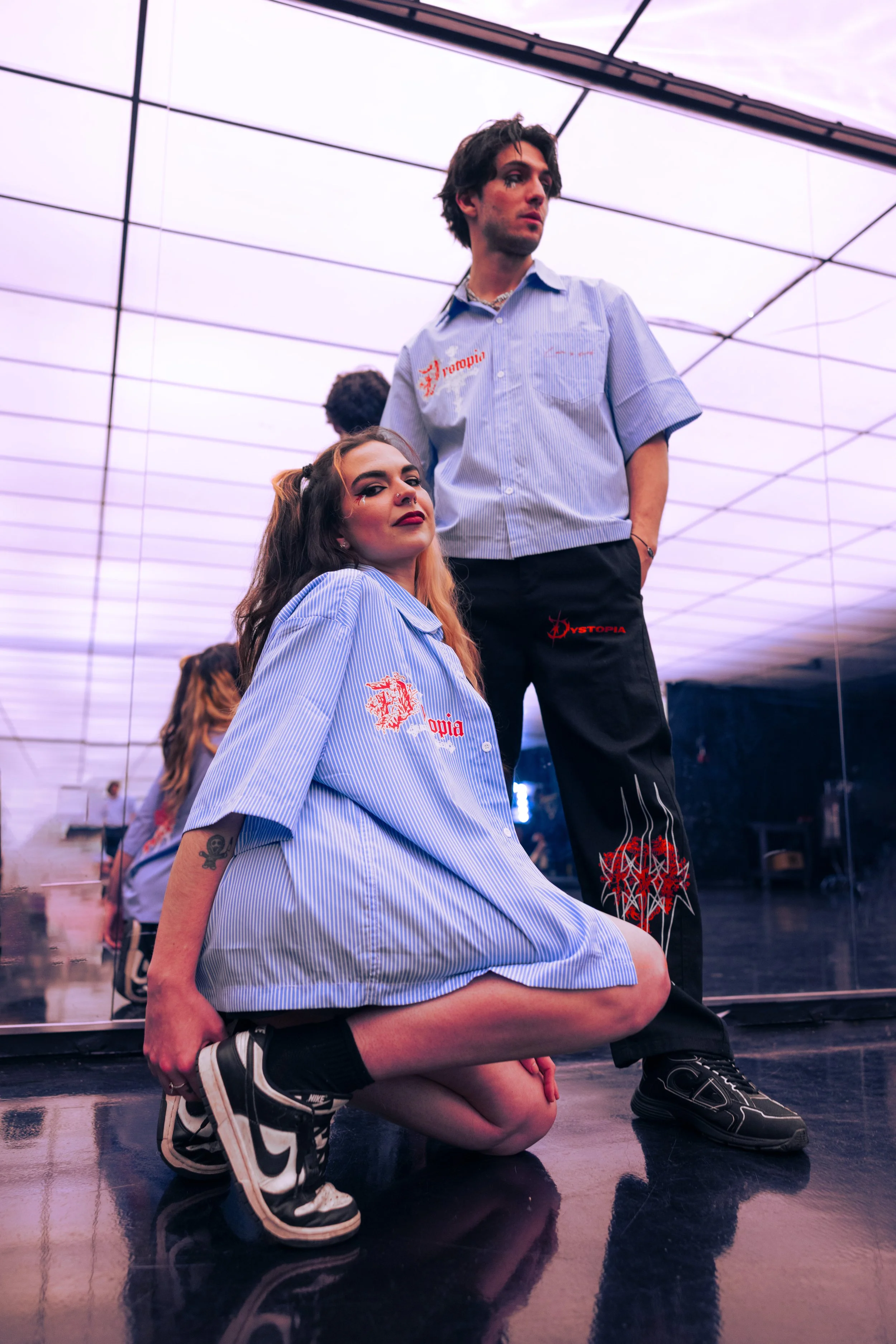 Two young adults, a woman kneeling and a man standing, pose in a room with a mirrored wall. They are wearing matching blue striped shirts with red embroidery that says 'Hystopia'. The woman has long, dark hair with orange highlights, and the man has 