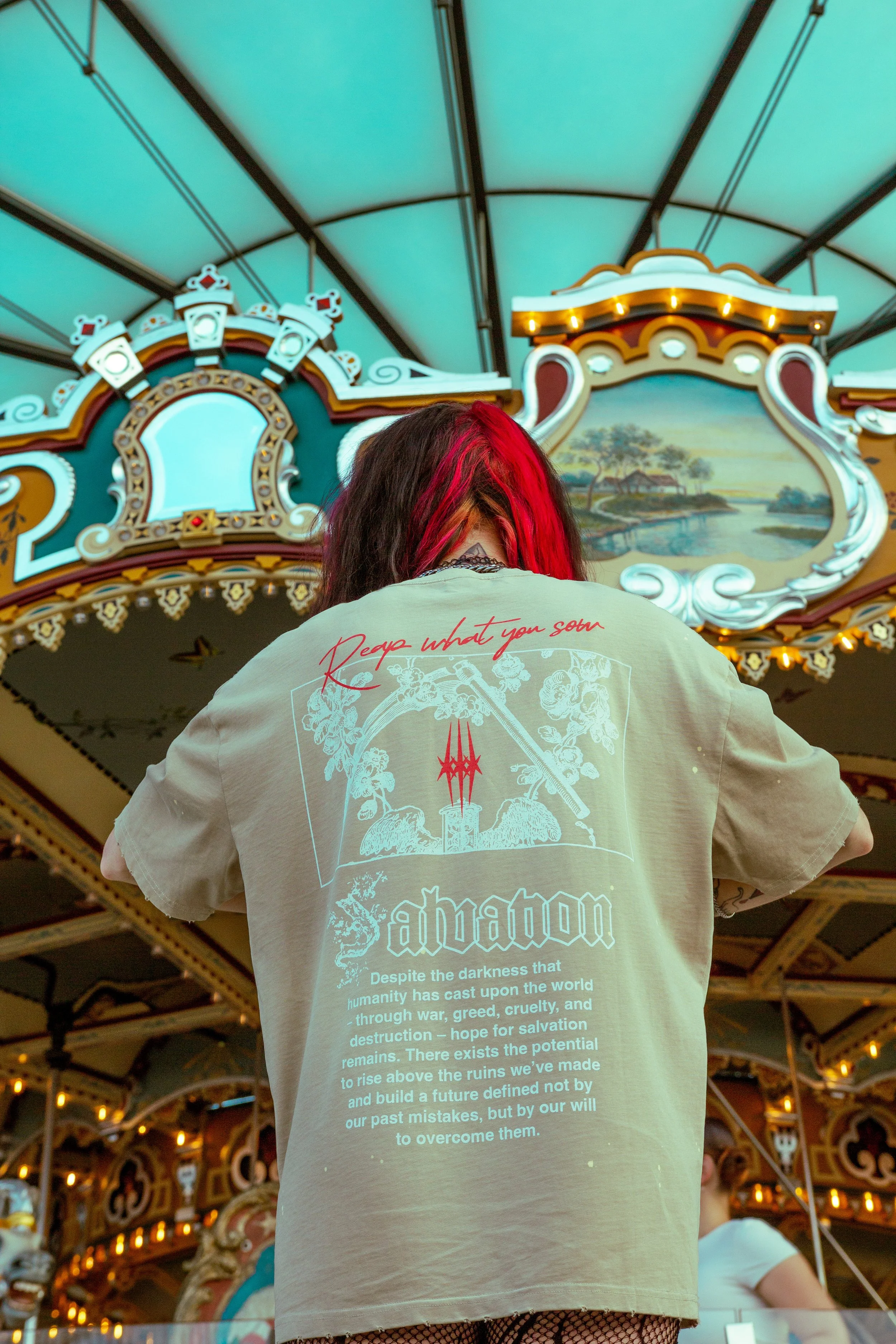 Person with red-dyed hair wearing a beige t-shirt with gothic-style text and artwork, standing in front of a vintage-style carousel with ornate decorations and a landscape painting.