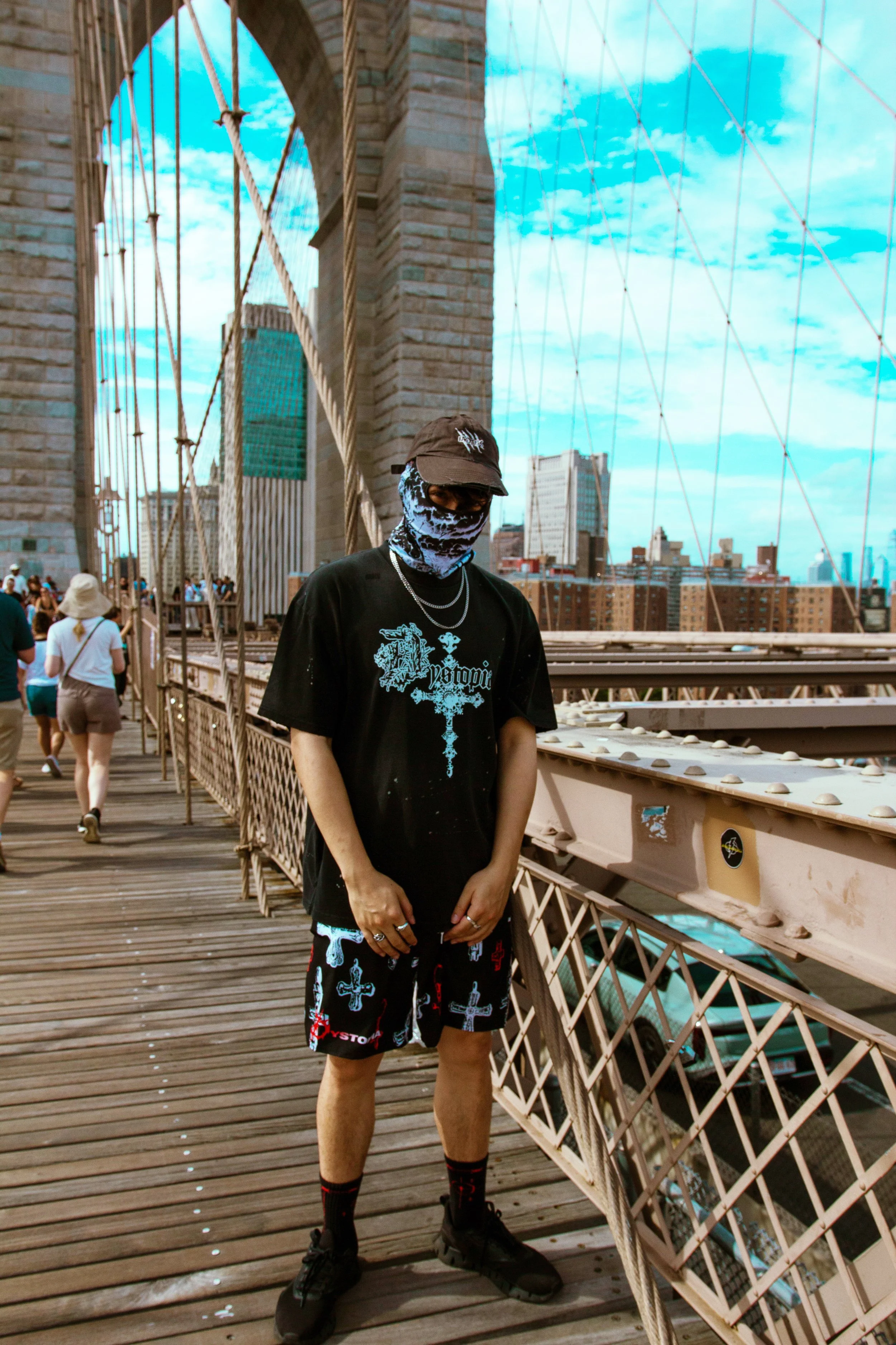 A person standing on a bridge in New York City, wearing a black t-shirt with a blue graphic and text, shorts with crosses, a mask, and sunglasses, with skyscrapers in the background.