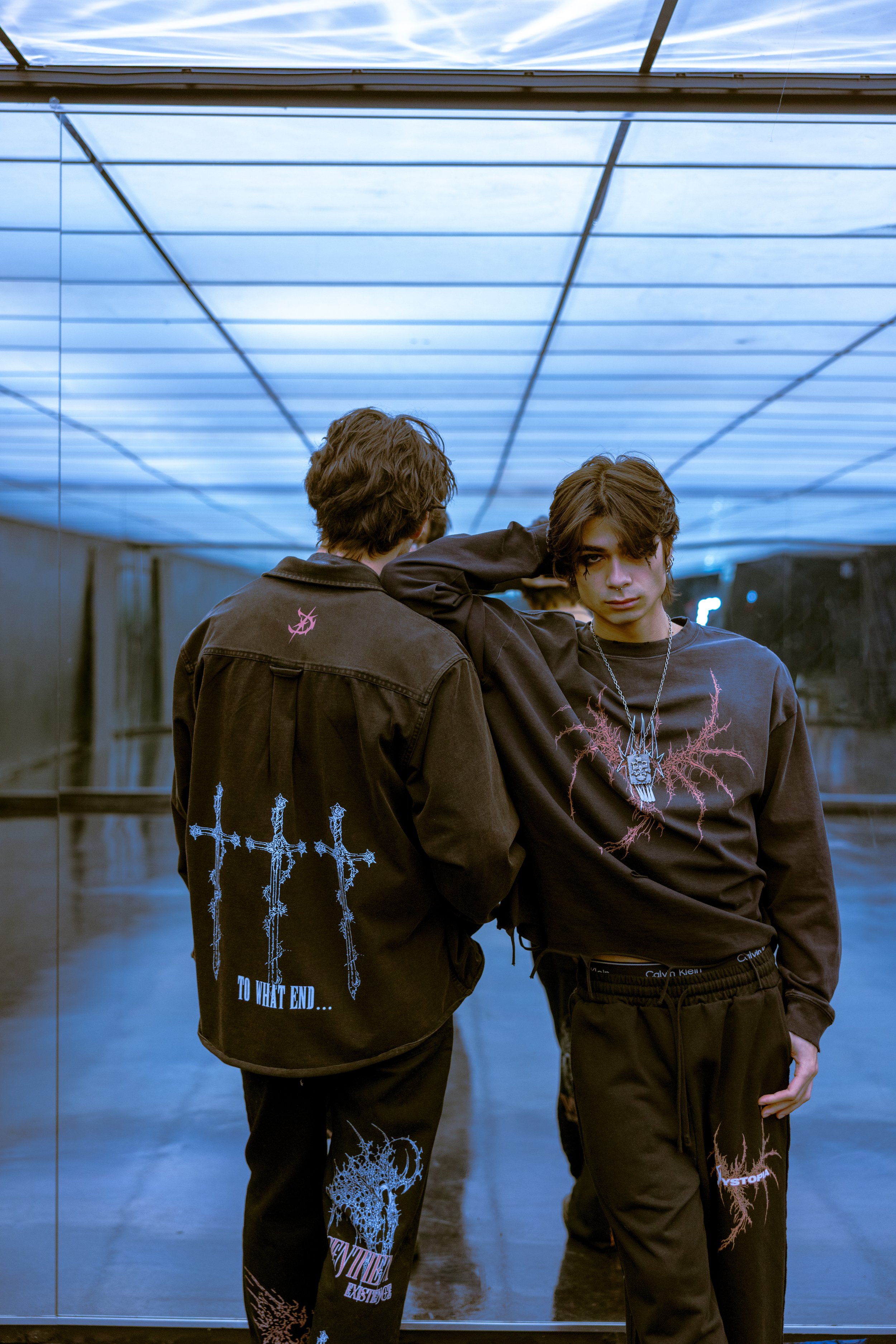 Two young men dressed in gothic or metal band style clothing, standing in front of a mirrored wall with a blue-lit ceiling. One faces away, showing back details, and the other faces forward, with one hand behind his head.