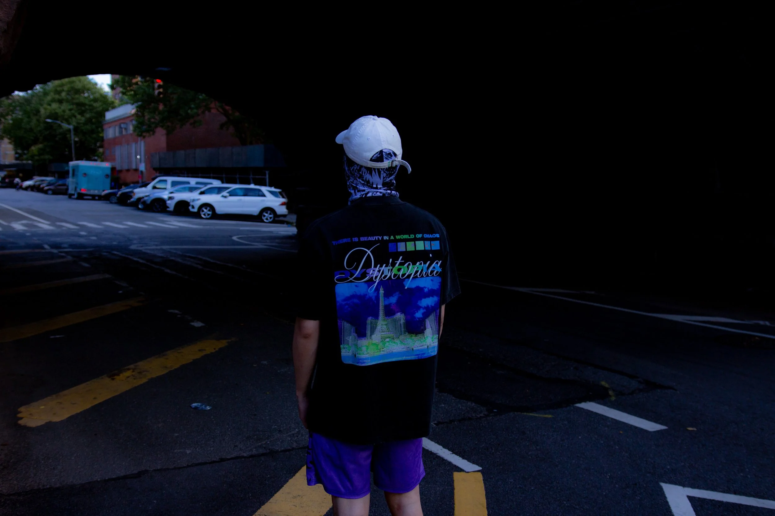 Person standing under a bridge in an urban environment, wearing a black jacket with a colorful cityscape and the word 'Aptopia' on the back, a white cap, purple shorts, and a face covering with a blue pattern.