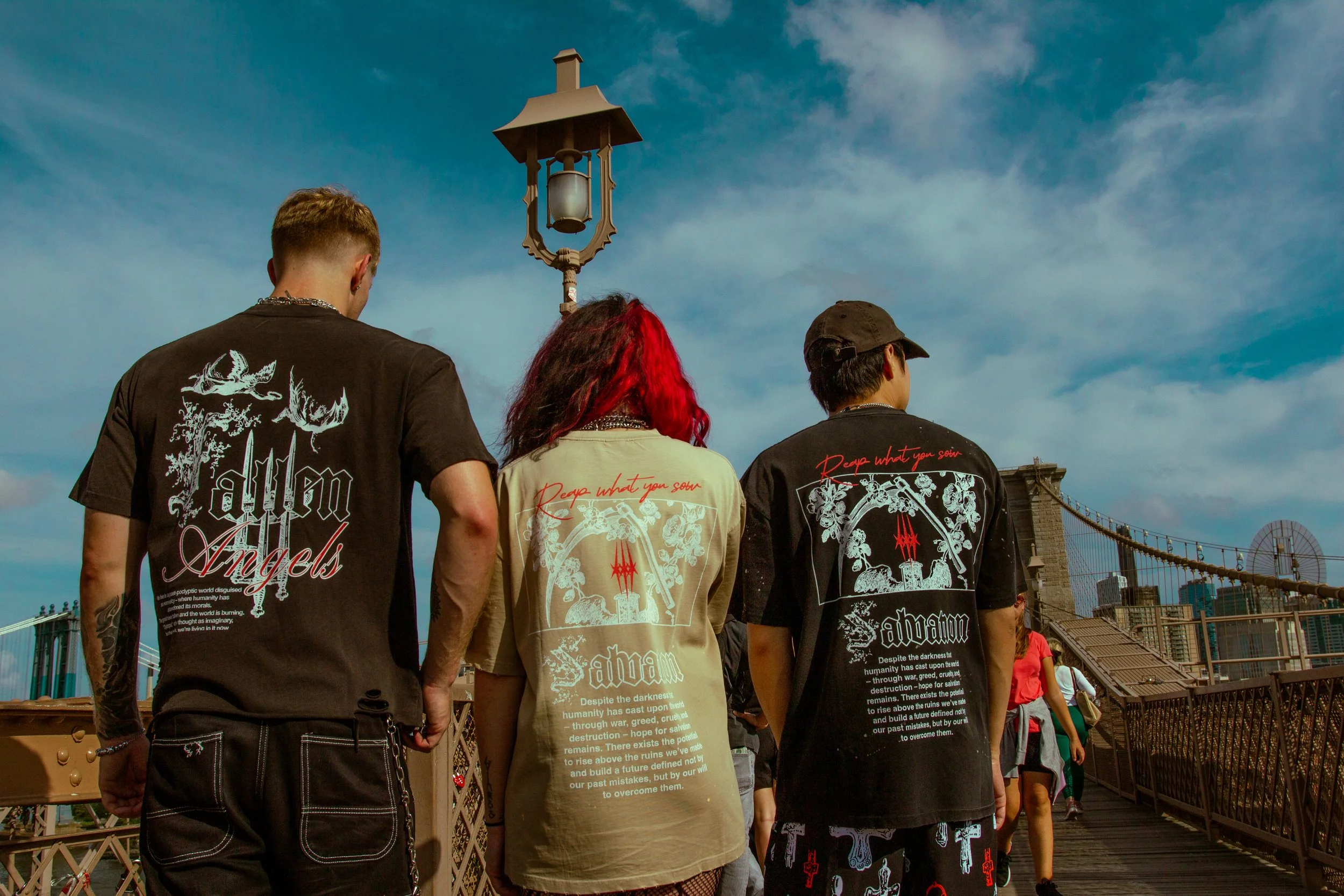 Three people with their backs turned, walking on a bridge against a blue sky with clouds. They are wearing dark and light printed t-shirts featuring artwork and phrases related to 'Alan Angels' and 'Absam,' with red and white graphic designs. The bri