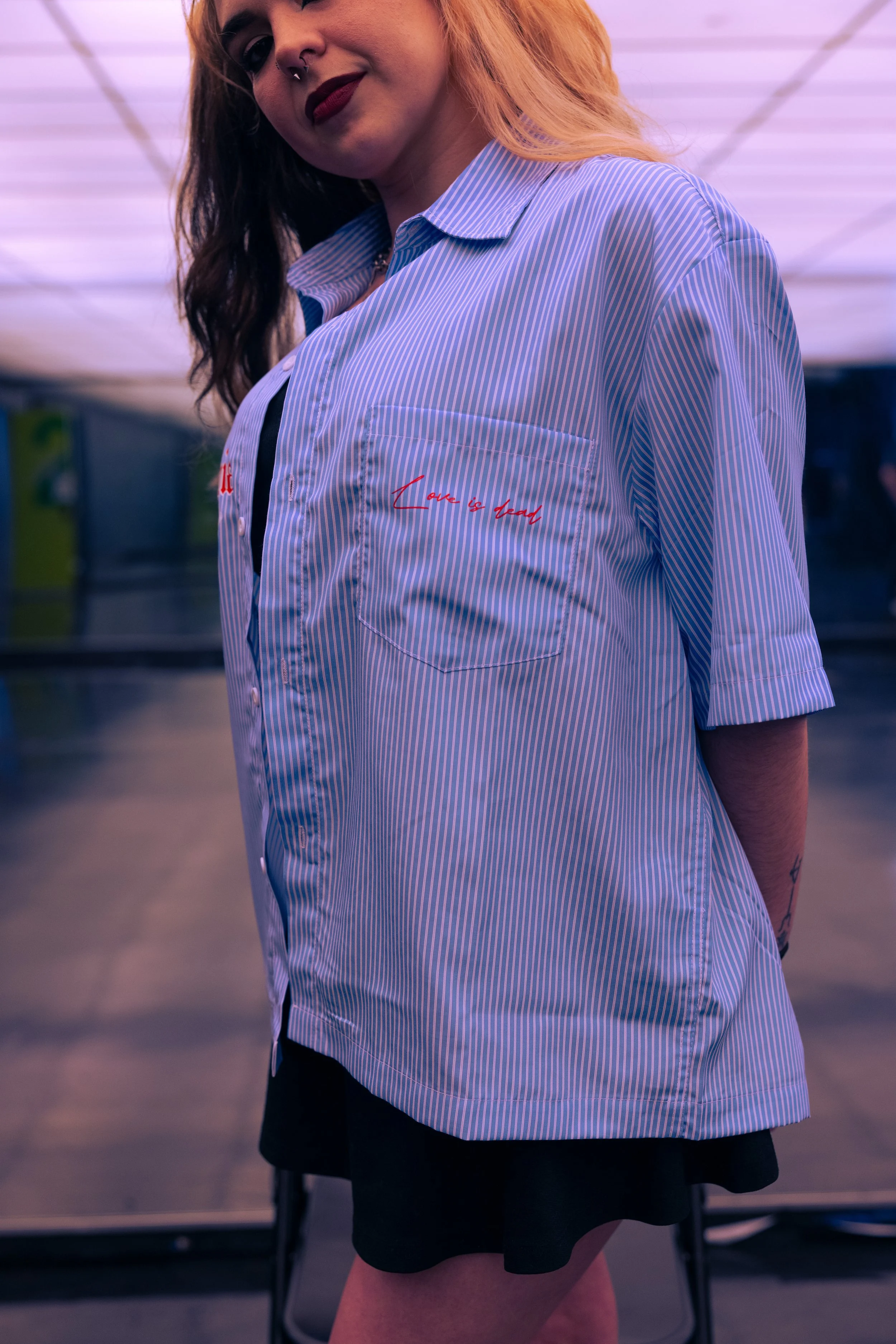 A young woman with wavy hair and a nose piercing, wearing a blue and white striped button-up shirt with red embroidery on the chest pocket that says "Love is dead," stands in an indoor setting with purple lighting, smiling slightly at the camera.