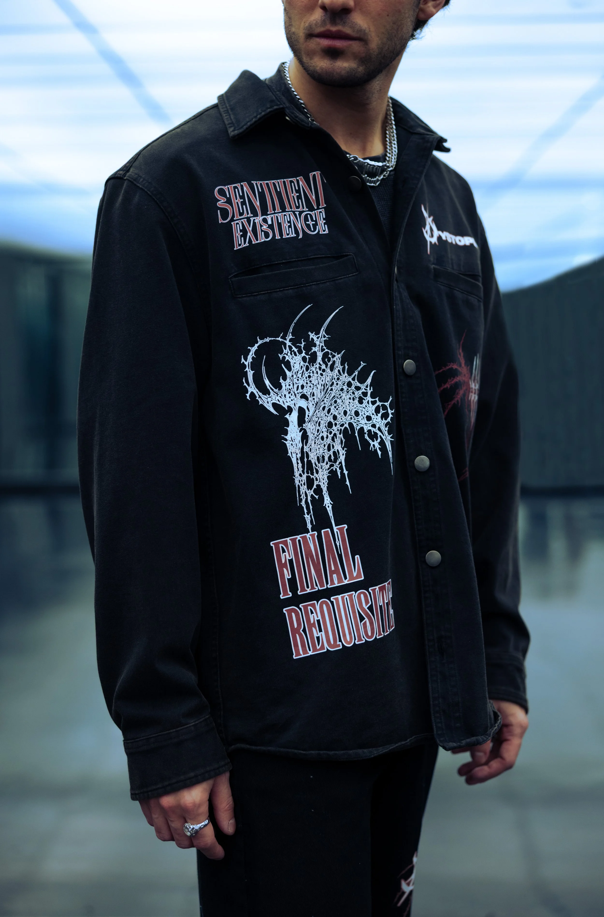 Close-up of a man wearing a black jacket with gothic-style text and graphics. The jacket features patches with words like 'Sentience' and 'Final Requiem' and detailed designs. The man is standing against a blurred outdoor background.