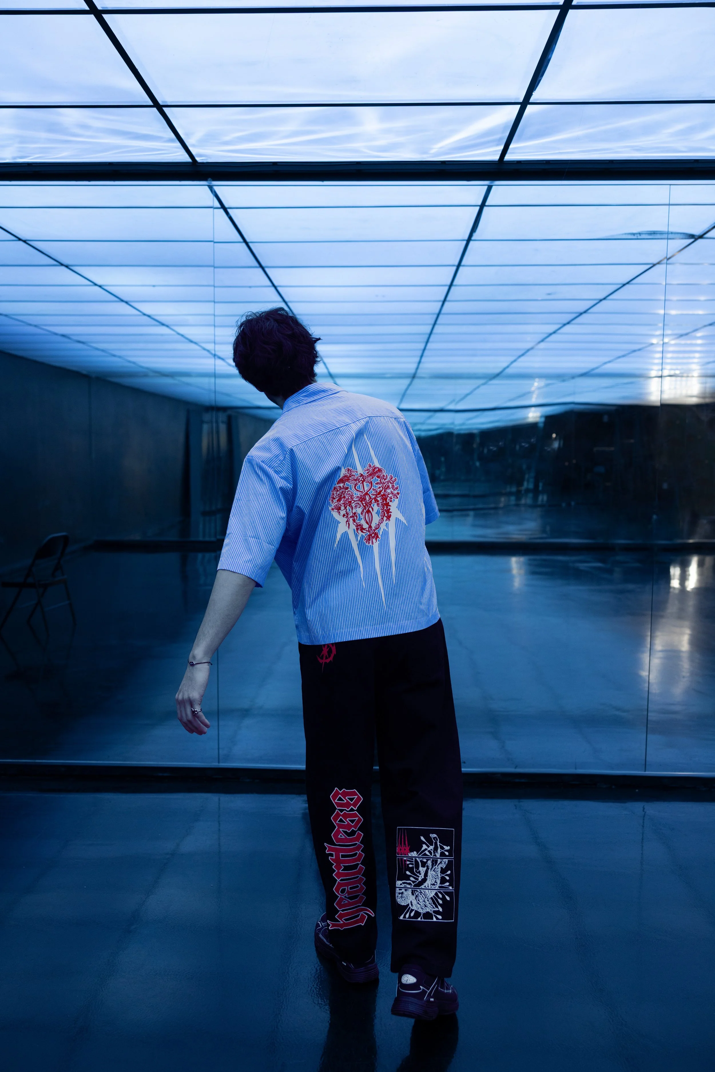 A person standing in a room with blue lighting and mirrored walls, wearing a light blue striped shirt with a red heart design on the back, black pants with red and white graphics, and sneakers, facing away from the camera.