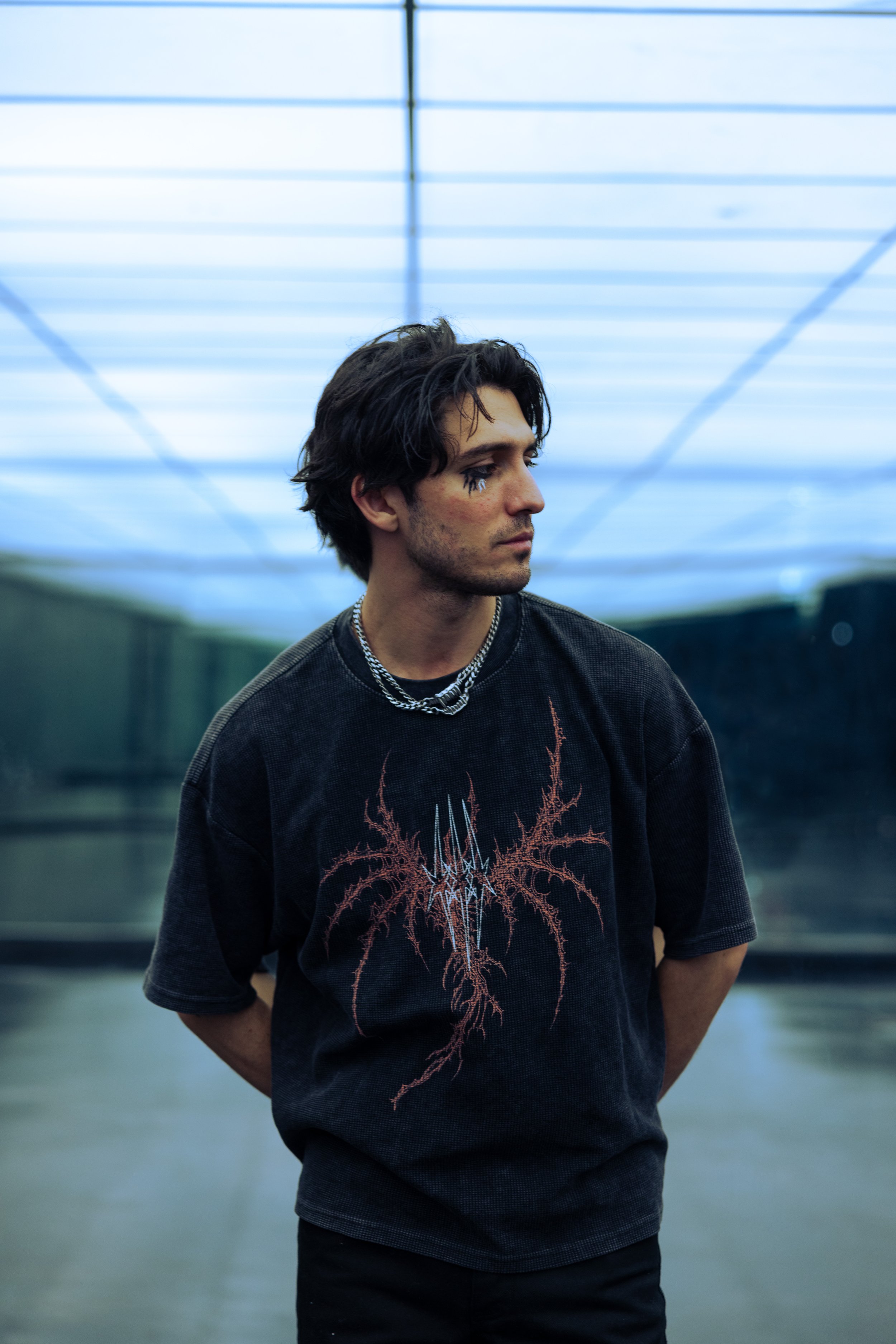 A young man with dark, messy hair and makeup around his eyes stands in a modern indoor space with a blue-lit ceiling and reflective walls, wearing a black T-shirt with a gothic design and layered silver necklaces.