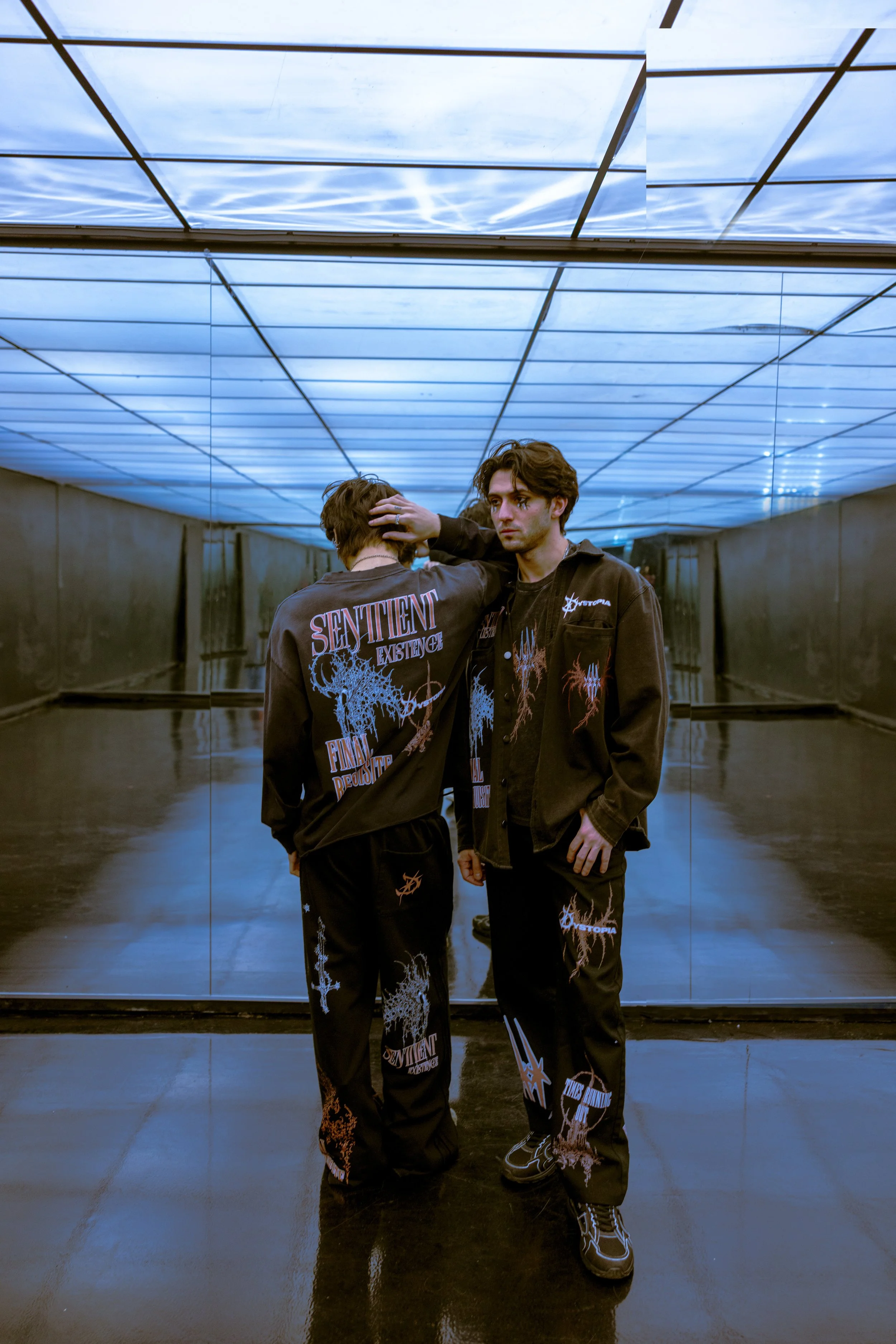 Two young men wearing black graphic clothing standing in front of a mirror in a room with a blue-lit ceiling.