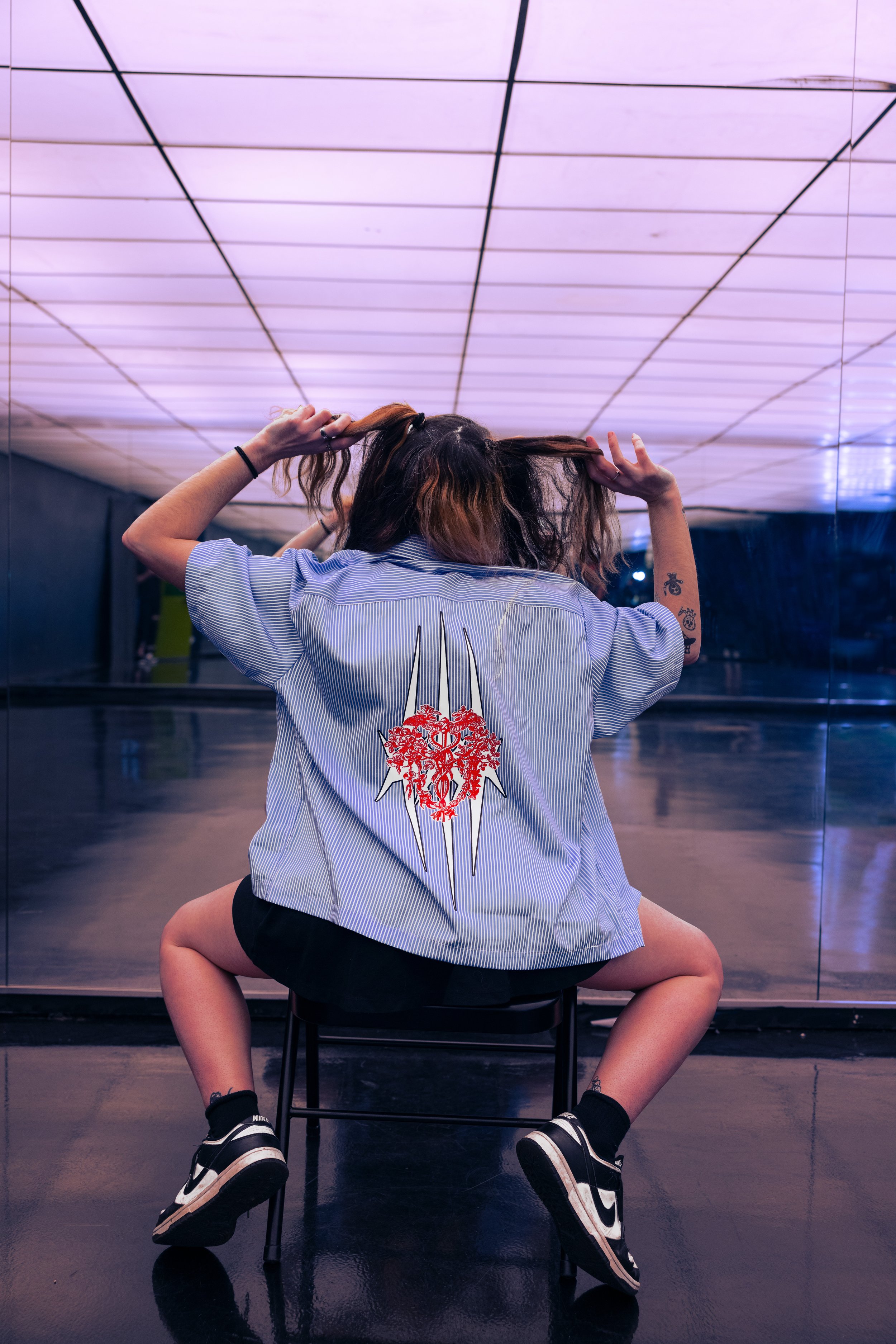 Person sitting on a chair with a mirrored wall behind them, wearing a loose striped shirt with a red heart and knife-shaped design on the back, black shorts, and black Nike sneakers. The person has tattoos on their arm and is holding their hair up.