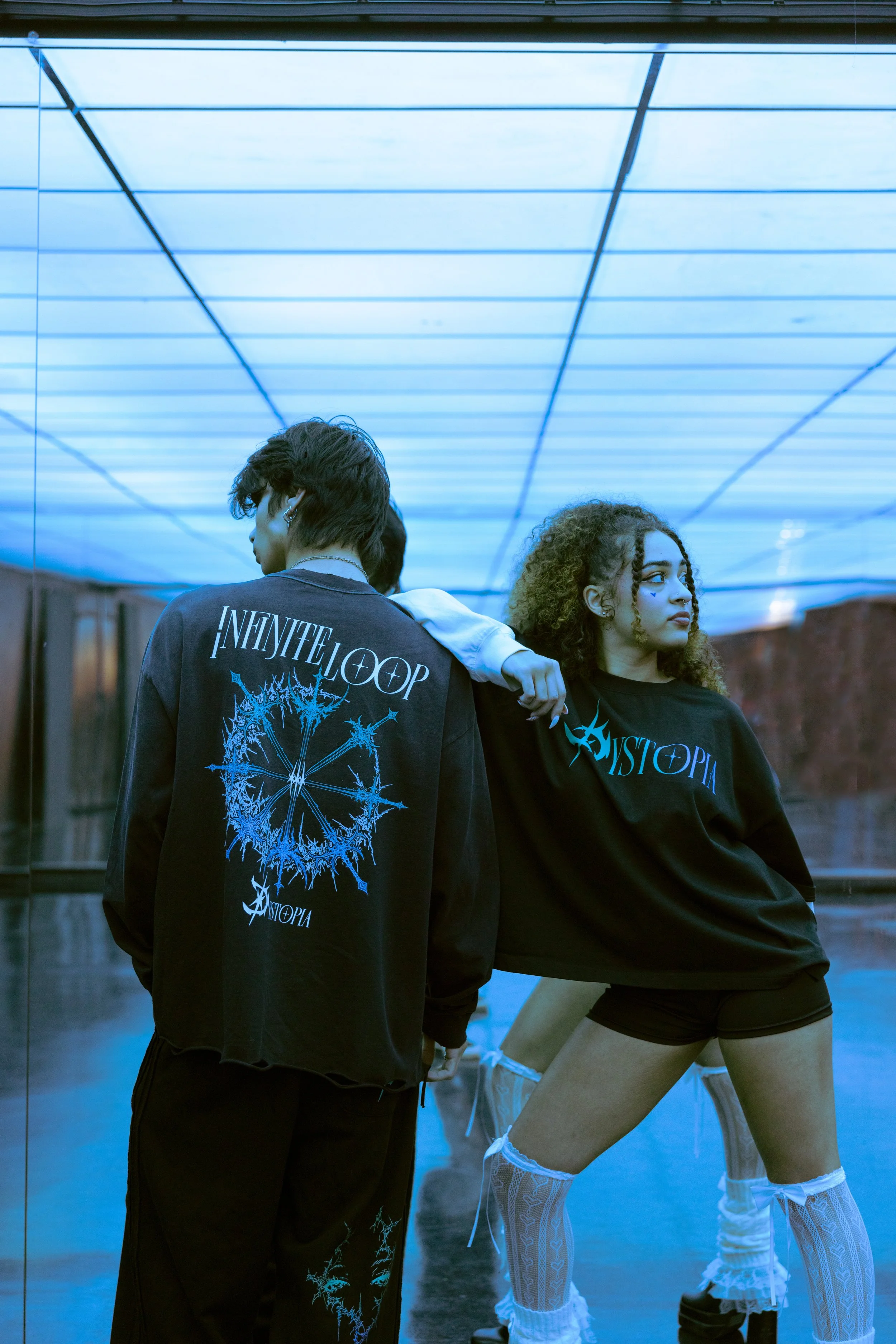Two young adults pose in front of a reflective surface, wearing black artistic fashion with glow-in-the-dark designs and text.