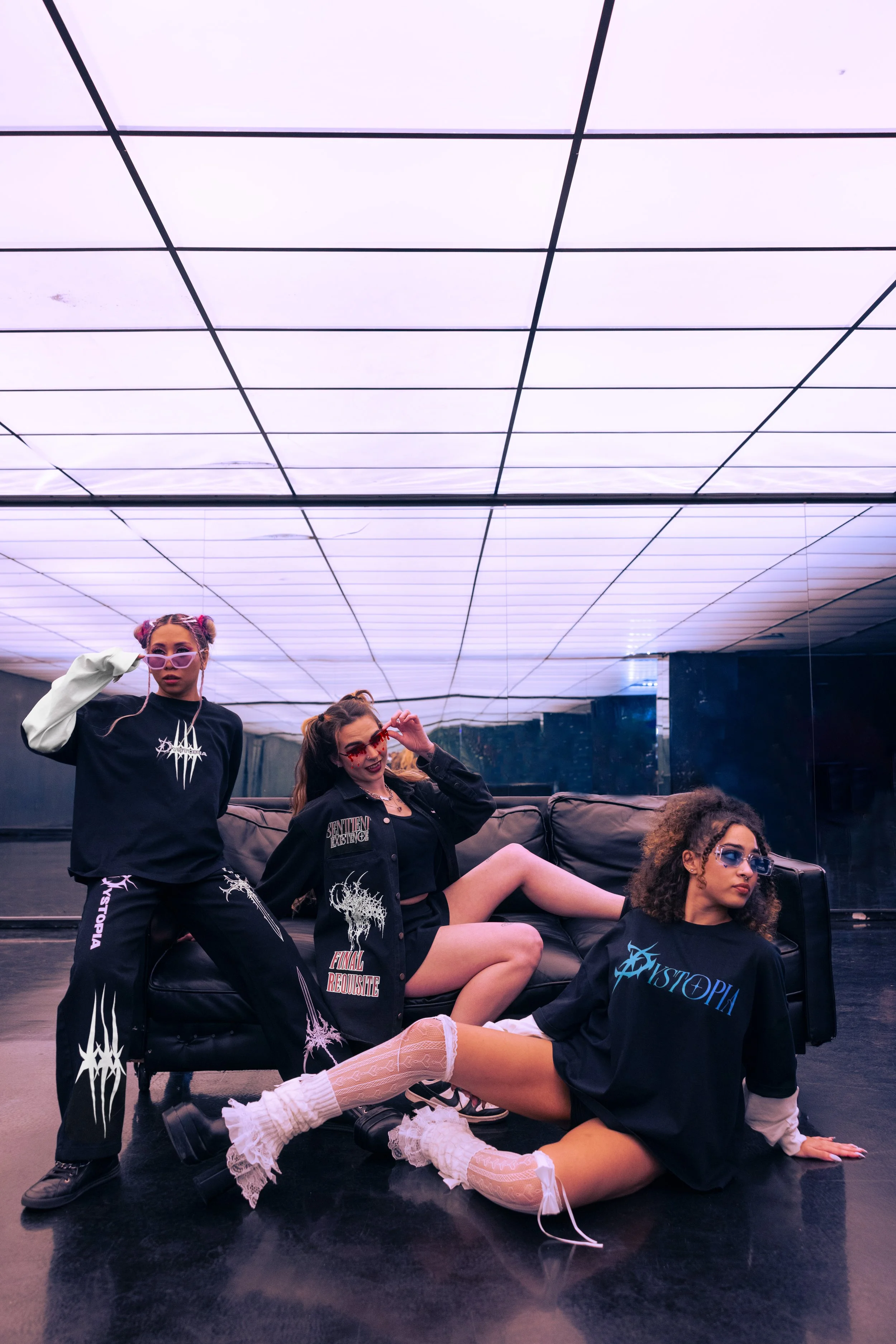 Three young women with colorful glasses and edgy clothing posing in front of a mirror on a black floor and illuminated ceiling.