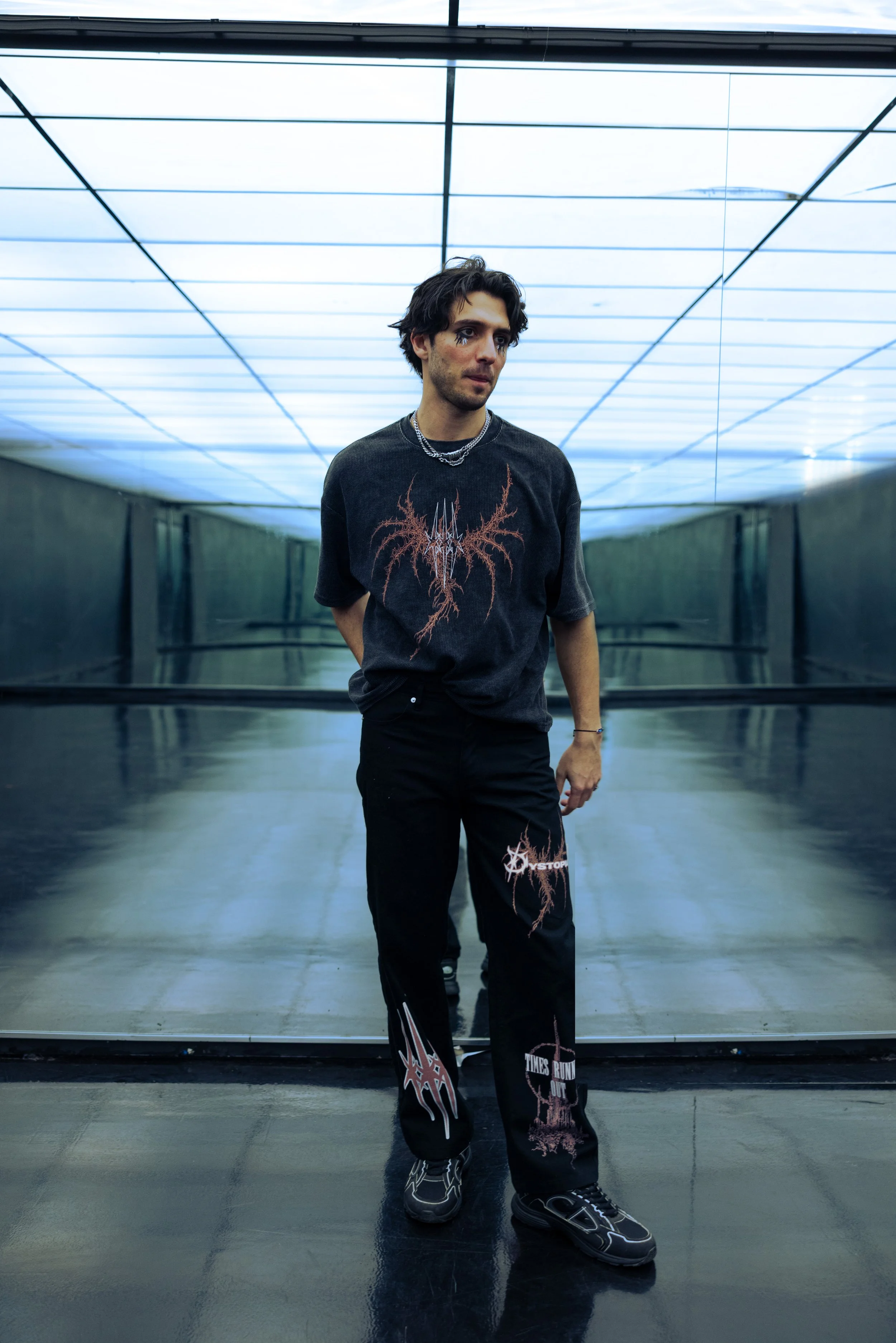 A young man with dark hair and light skin standing in front of a mirrored wall in a modern, illuminated setting, wearing black graphic t-shirt and pants with various designs and text, along with sneakers and jewelry.