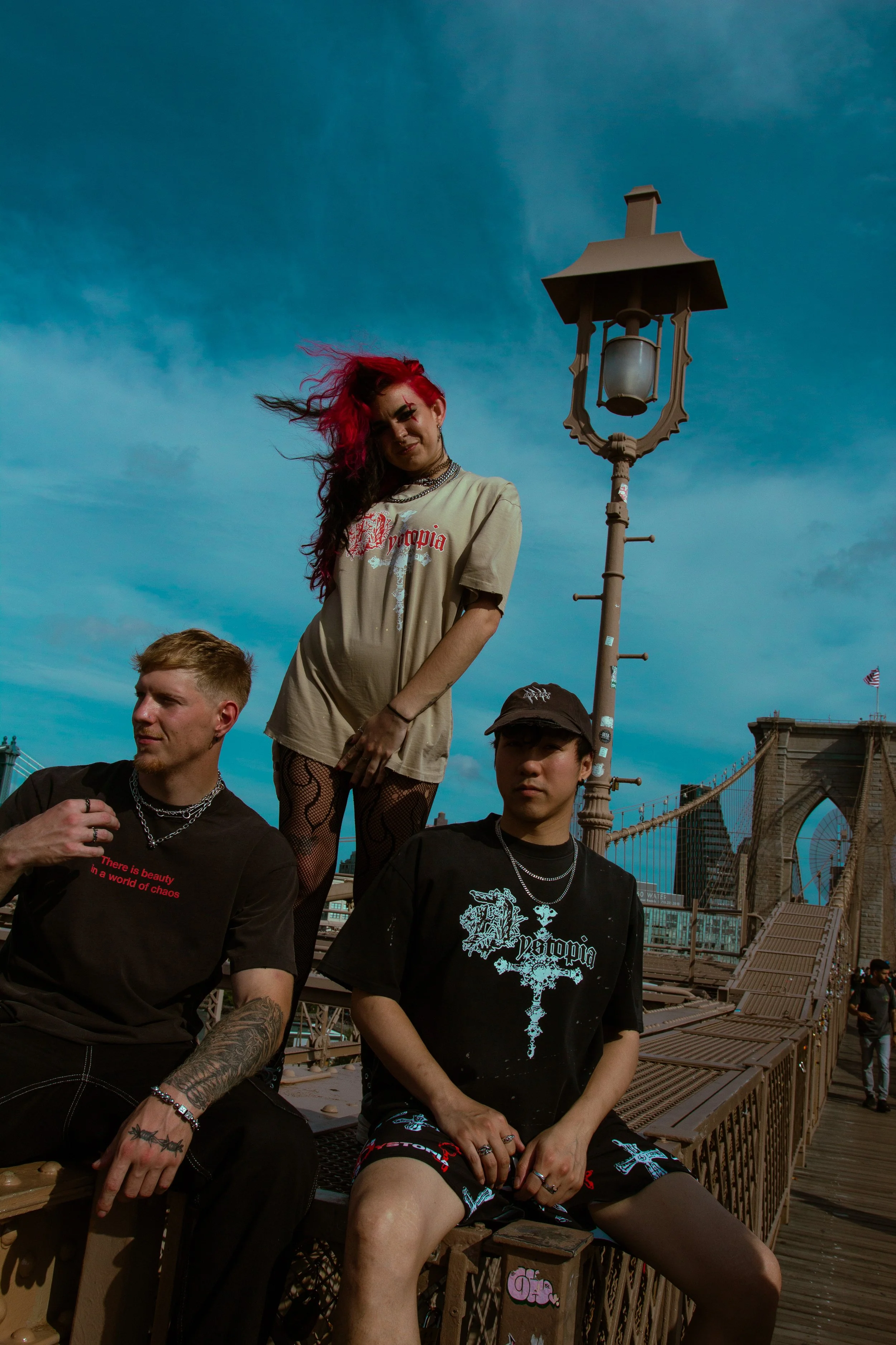 Three young people with alternative fashion styles sitting and standing on the Brooklyn Bridge in New York City, with the bridge and skyline in the background.