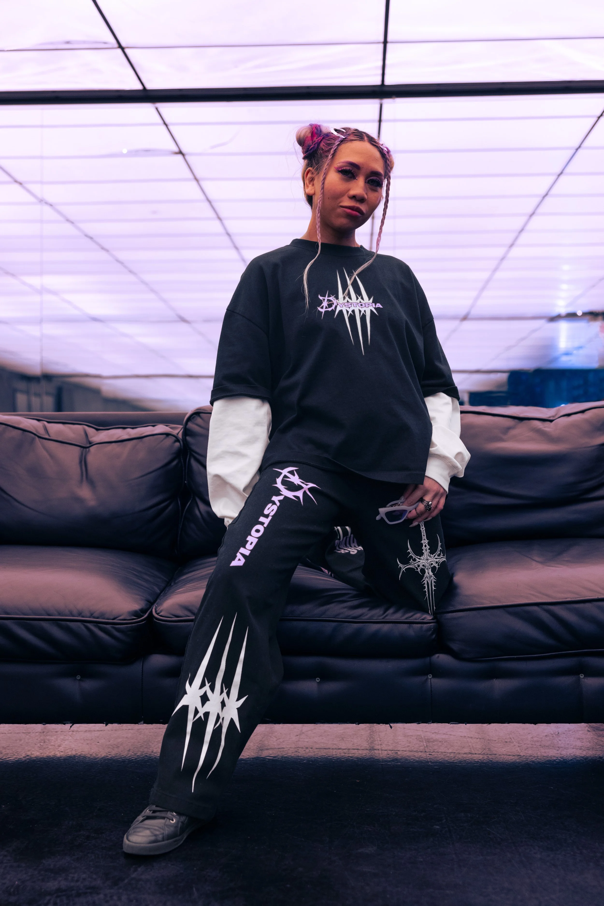 A young woman with pink braids wearing a black and white oversized hoodie and black pants with white graphics, standing in front of a sofa in a room with purple-lit ceiling panels.