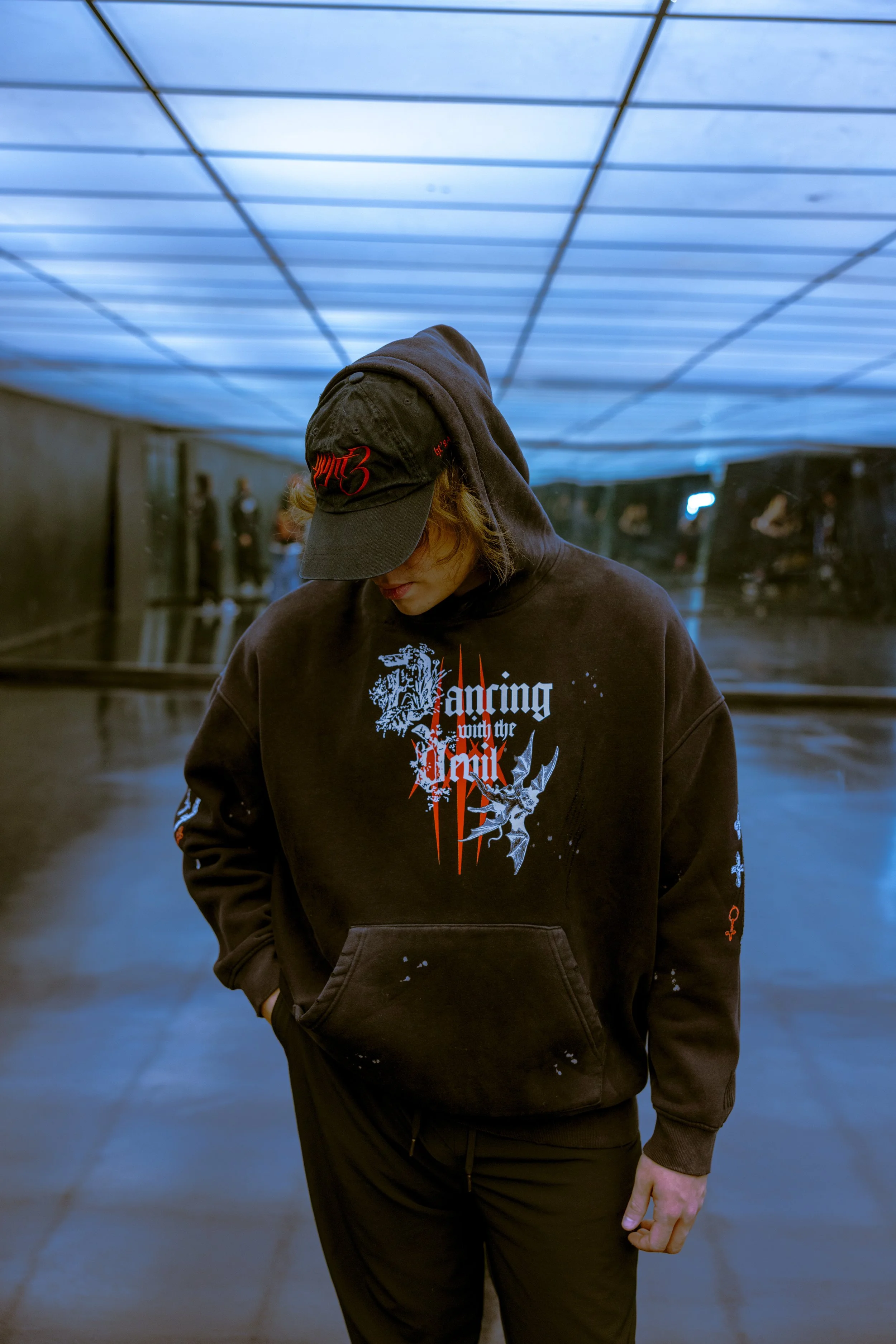 A person with blond hair wearing a black hoodie with graphics and text, and a cap with red lettering, standing in an underground tunnel with blue lighting and other people in the background.