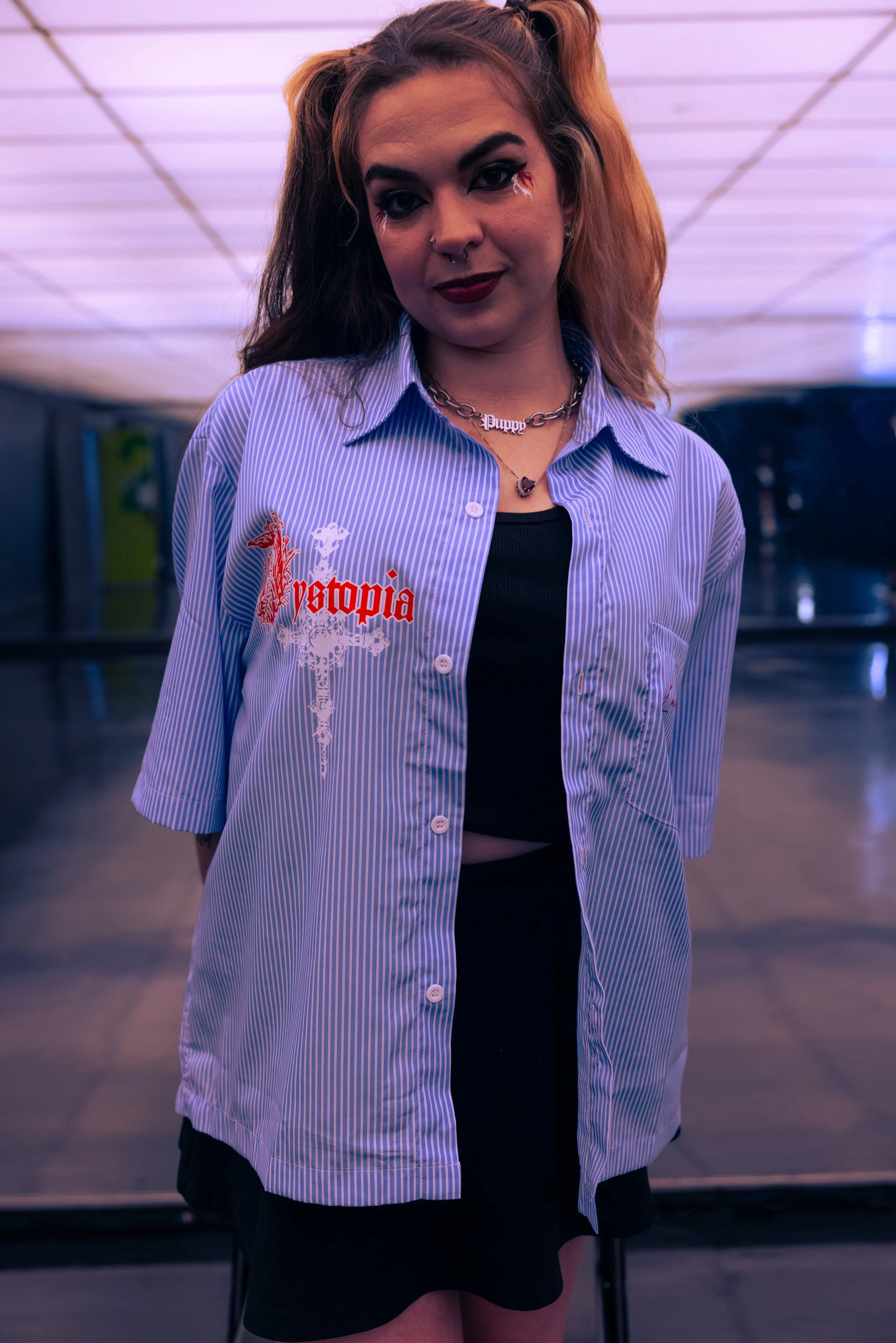 A young woman with layered hair, one side blond and the other dark, stands in front of a dark background. She is wearing a blue and white striped shirt with the word "Postopia" and a dragon design on it, over a black top and black skirt. She has make