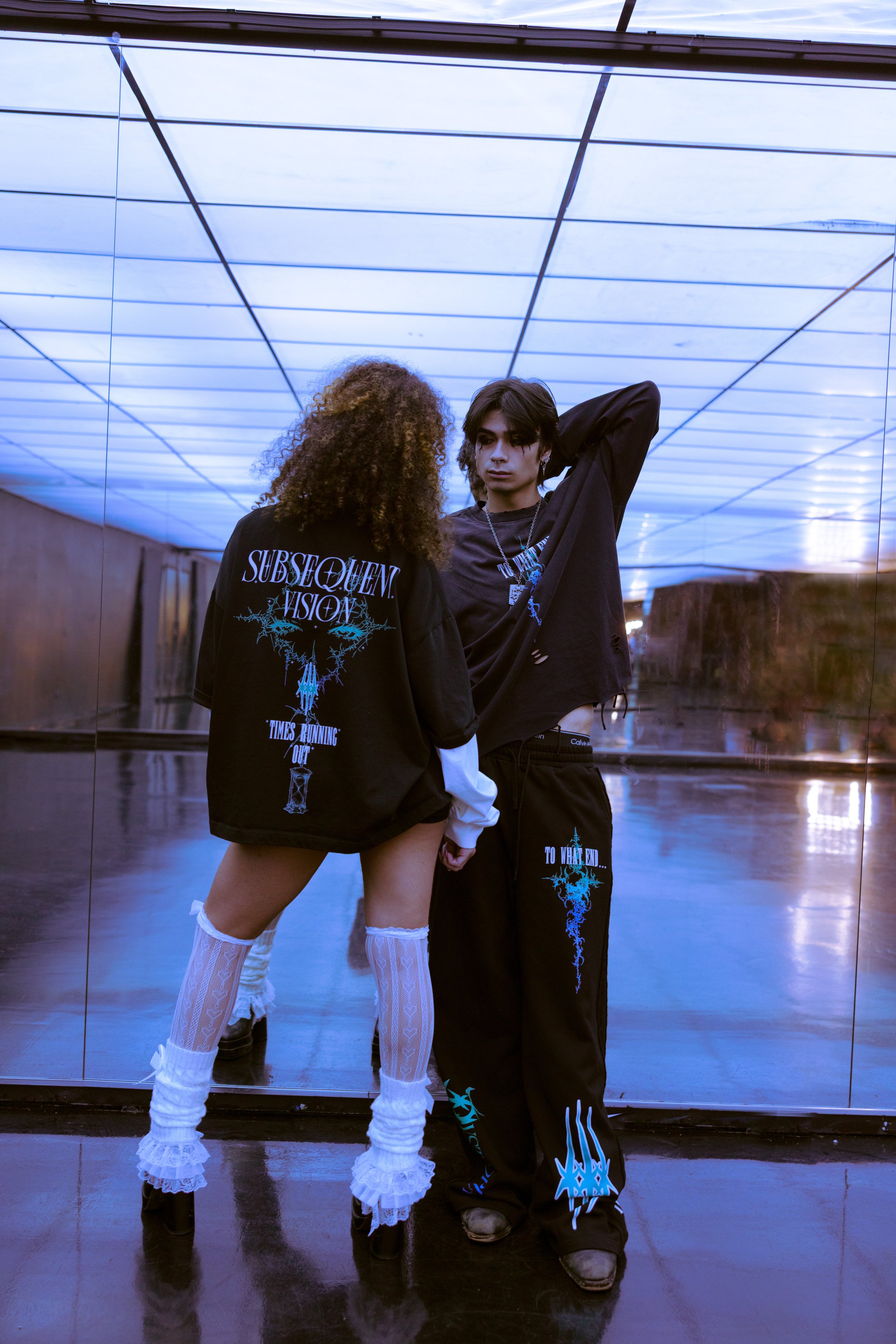 Two young adults posing in front of a mirrored wall under a blue-lit ceiling; the woman is wearing fishnet thigh-high socks and black shoes, and the man is wearing black pants and a black shirt with graphic designs.
