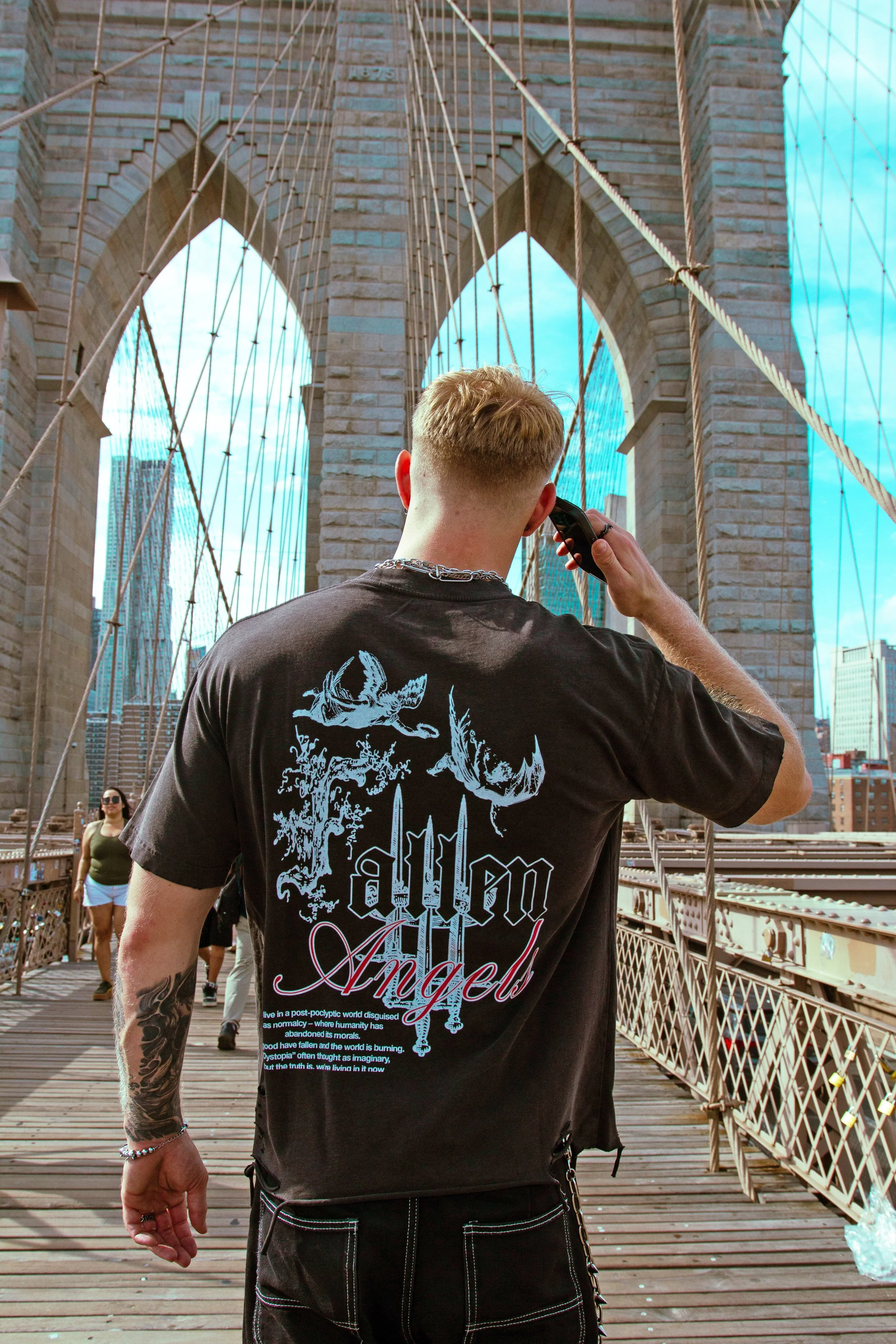 A man talking on a cellphone on the Brooklyn Bridge with a woman walking behind him and skyscrapers in the background.