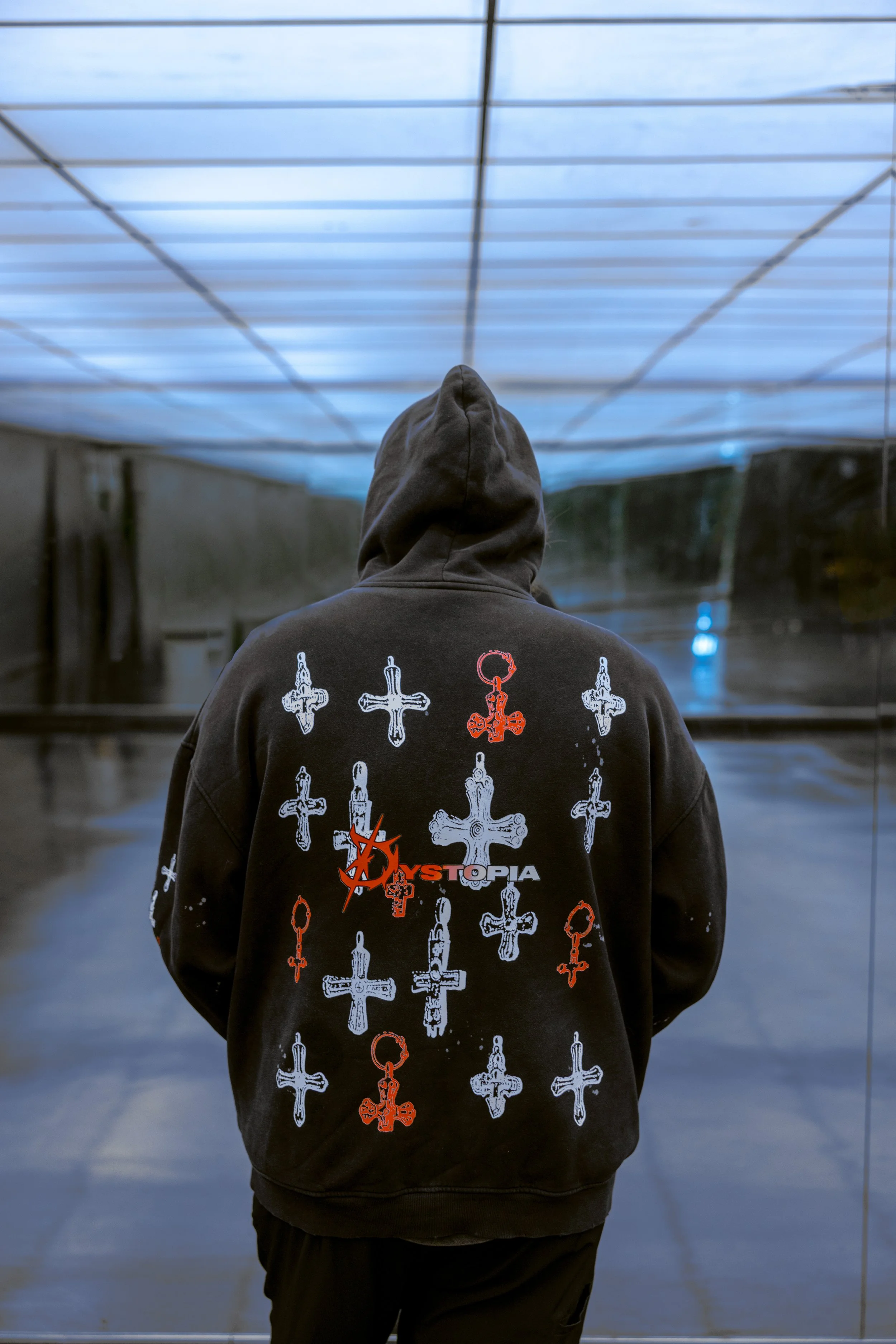 Person in a black hoodie with graphic crosses and the word 'HYSTOPIA' on the back, standing indoors with a reflective glass wall behind them.