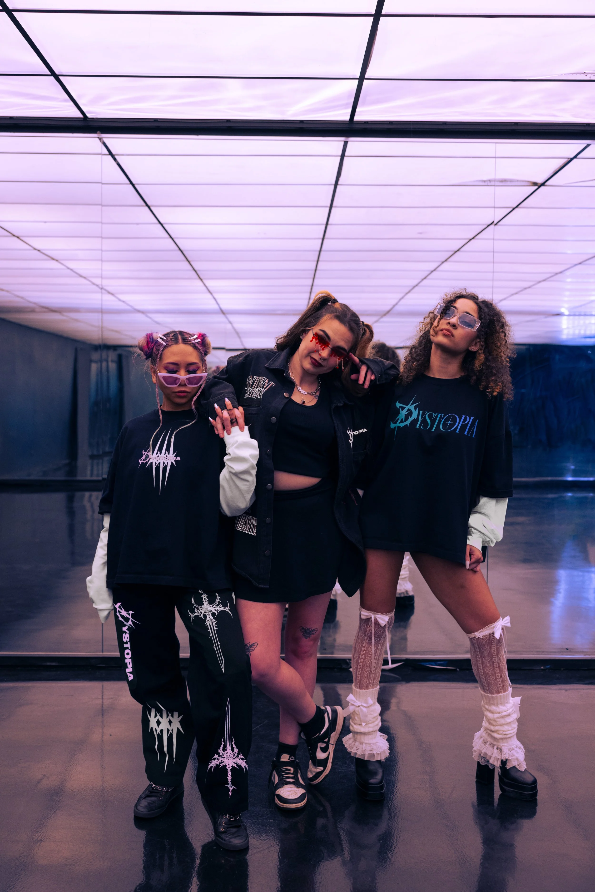 Three women in edgy fashion posing in front of a mirrored wall with purple ceiling lights.