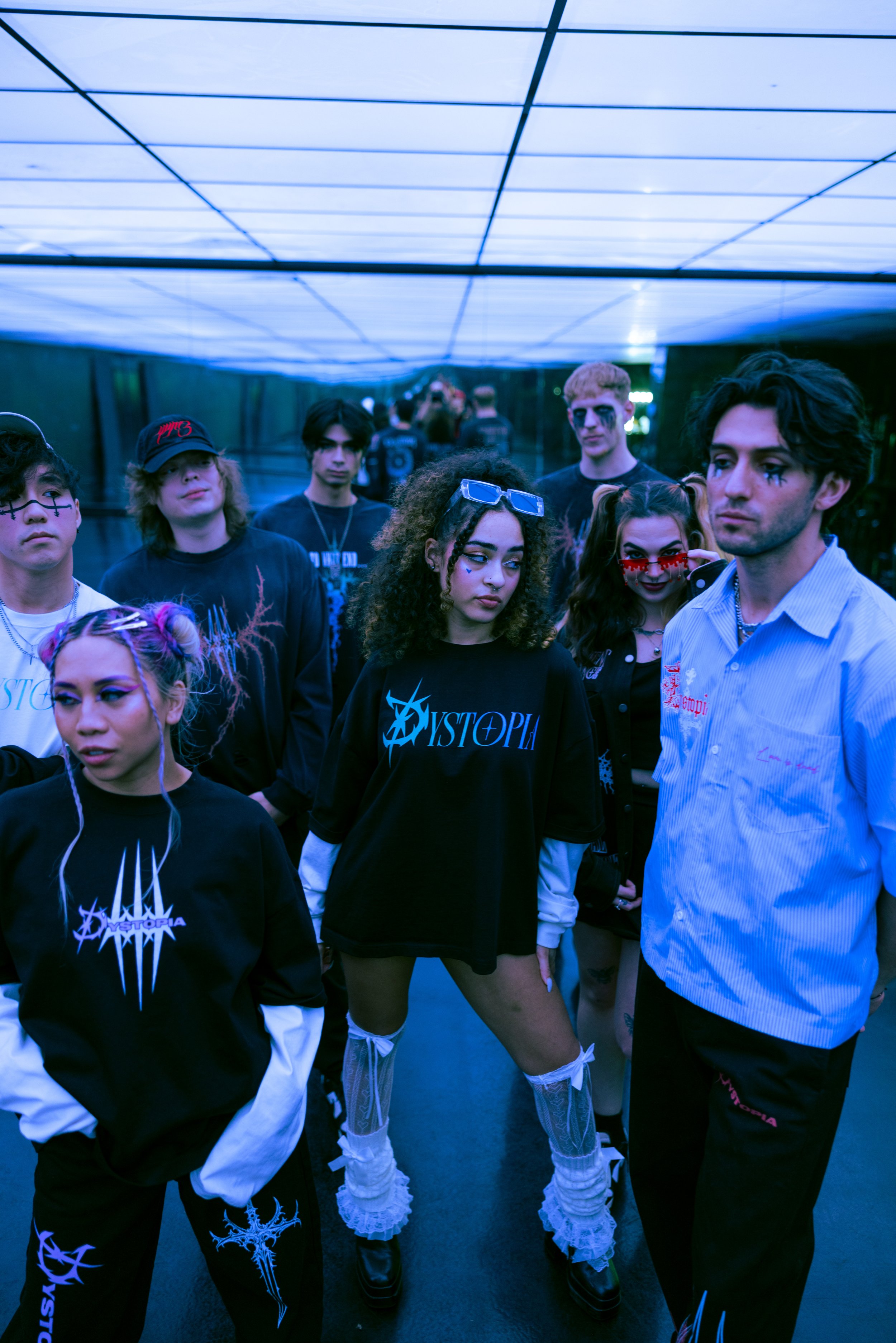 A group of young people dressed in alternative fashion, standing in a dimly lit, blue-toned indoor space with mirrored walls and ceiling lights.