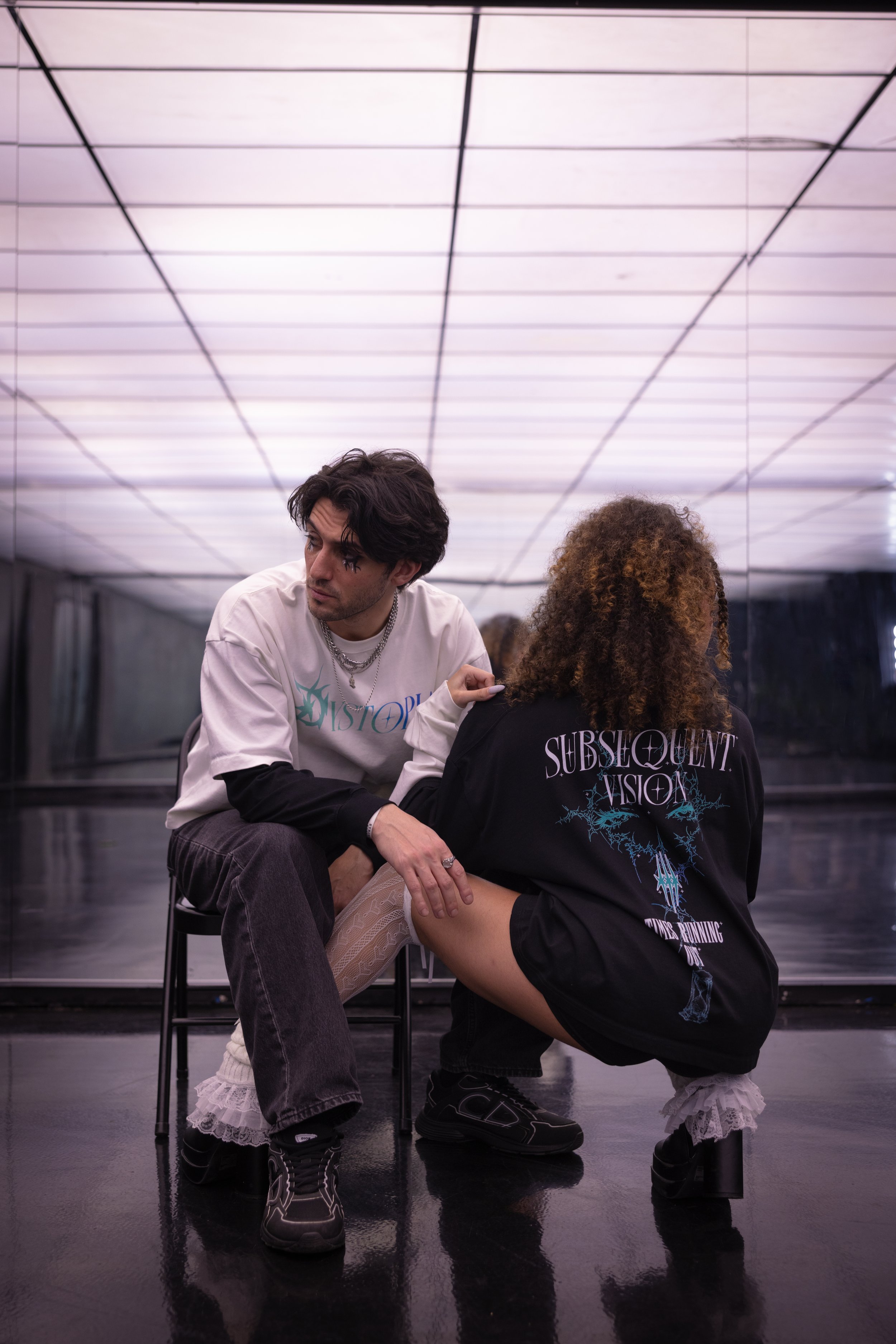Two people in a modern interior with reflective floors and a brightly lit ceiling, one sitting on a chair and the other crouching, both dressed in casual streetwear with graphic designs on their clothing.