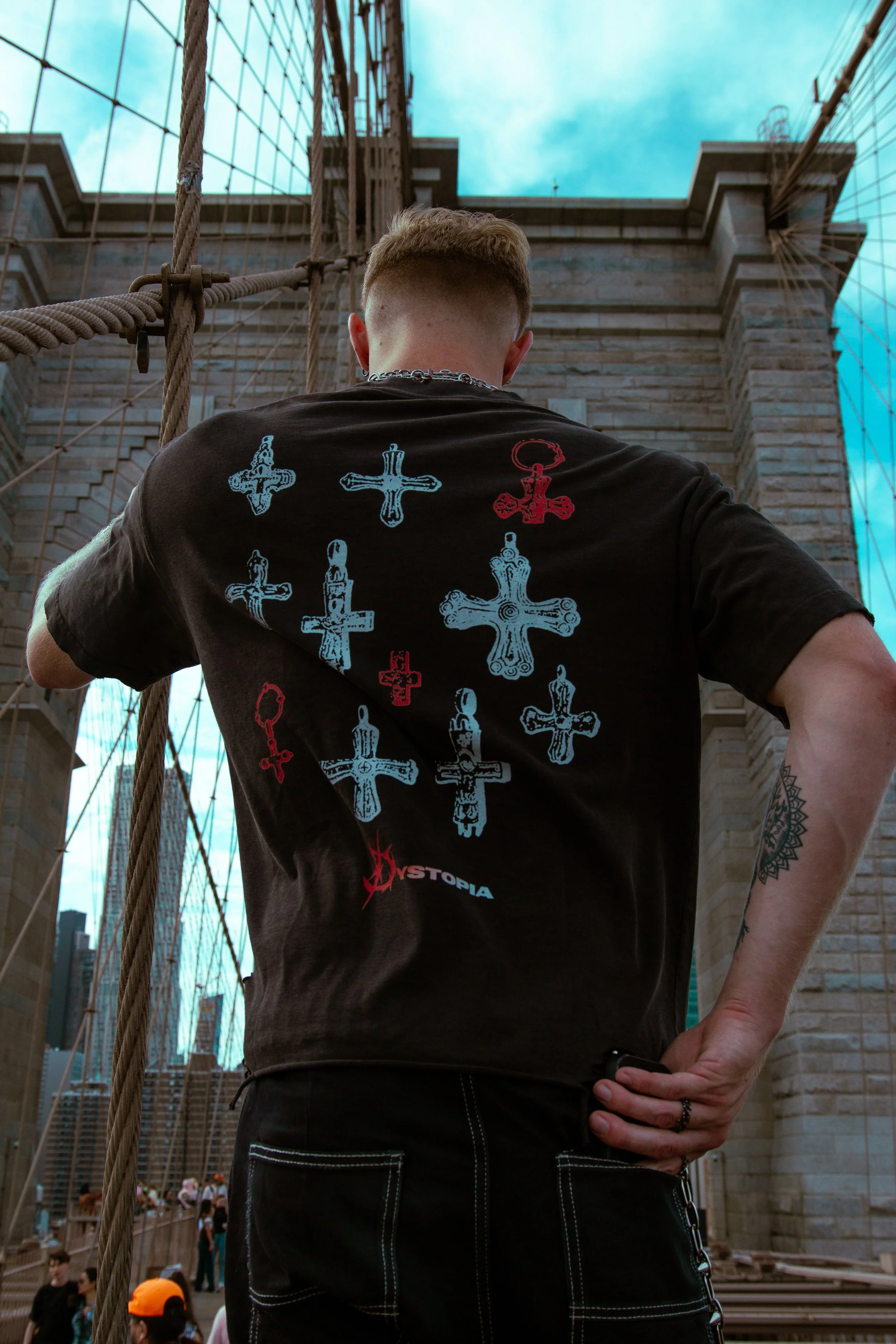 A man stands on a bridge with his back to the camera, wearing a black T-shirt decorated with various crosses and the word 'Utopia'. The bridge is part of the Brooklyn Bridge with its cables and stone towers visible, under a cloudy sky.
