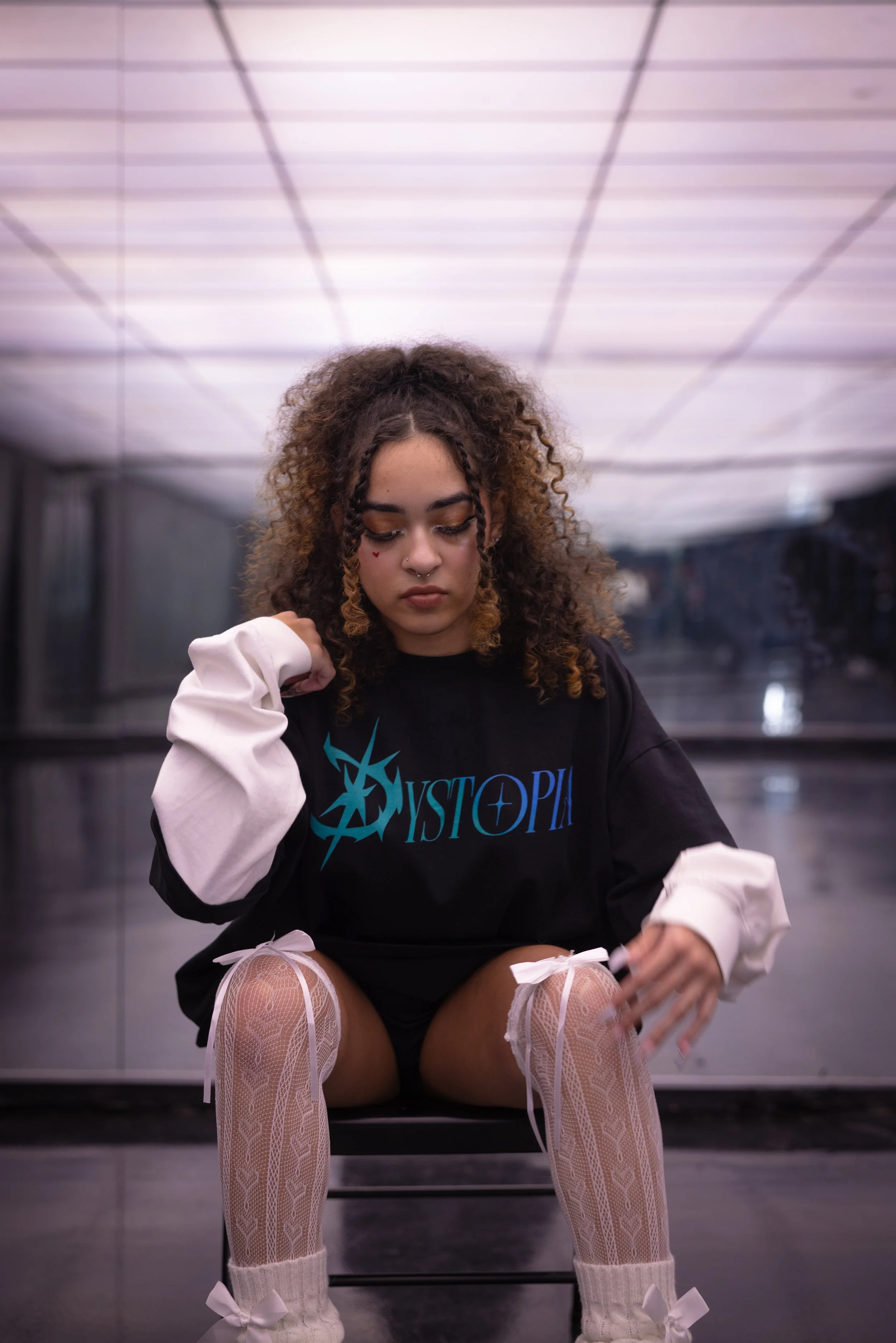A young woman with curly hair, dressed in a black and white oversized shirt with 'Mystopia' written on it, white lace tights with bows, and white ankle socks, sitting on a bench in an indoor setting with a reflective surface and a lit ceiling.