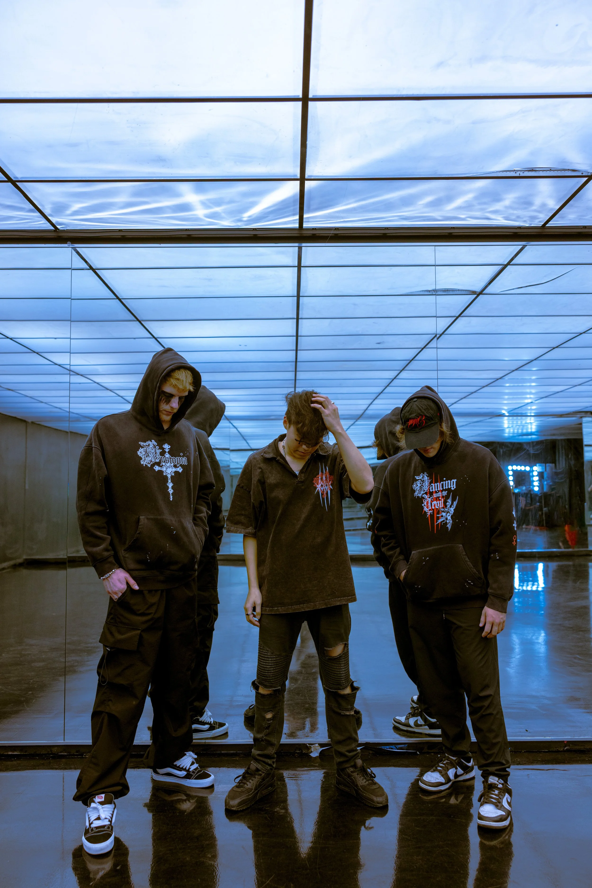 Three young men standing in front of a large mirror with a blue lit ceiling, dressed in dark streetwear, with one touching his head and the others looking down or away.