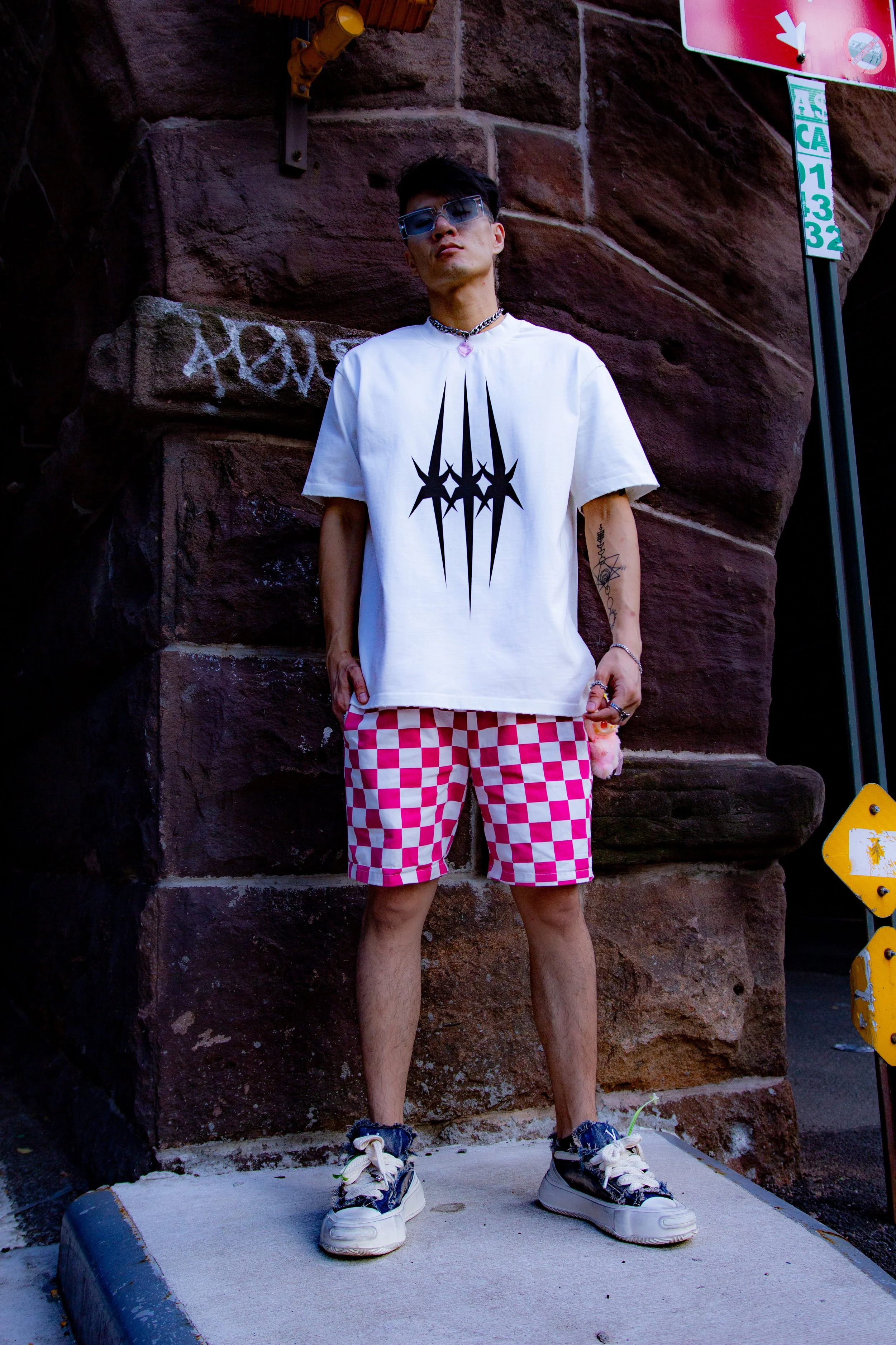 A person standing outdoors against a brick wall wearing sunglasses, a white graphic t-shirt, pink and white checkered shorts, and sneakers.