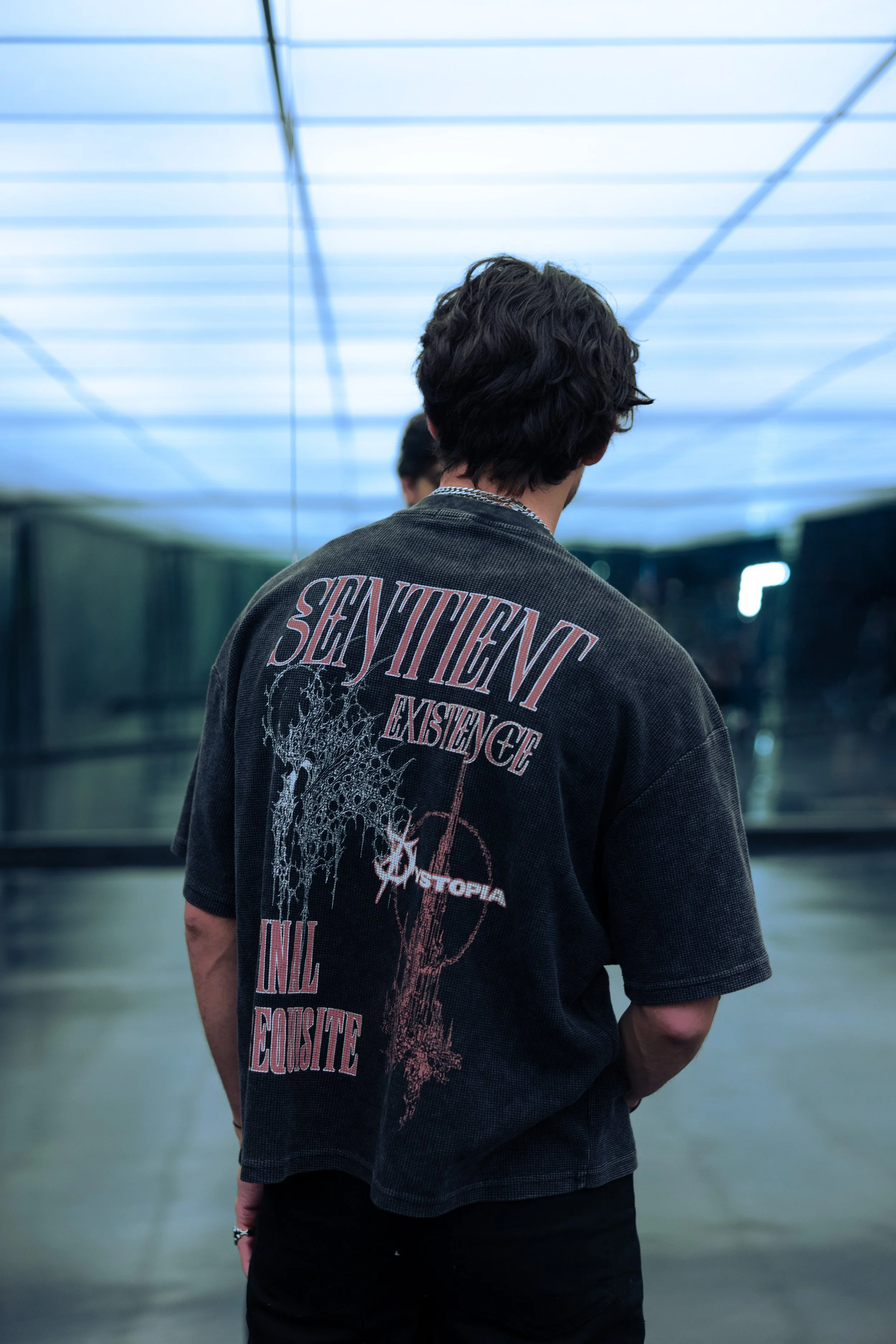 A young person with curly dark hair viewed from behind, wearing a black graphic T-shirt, standing in an indoor space with a glowing blue-lit ceiling and mirrors.