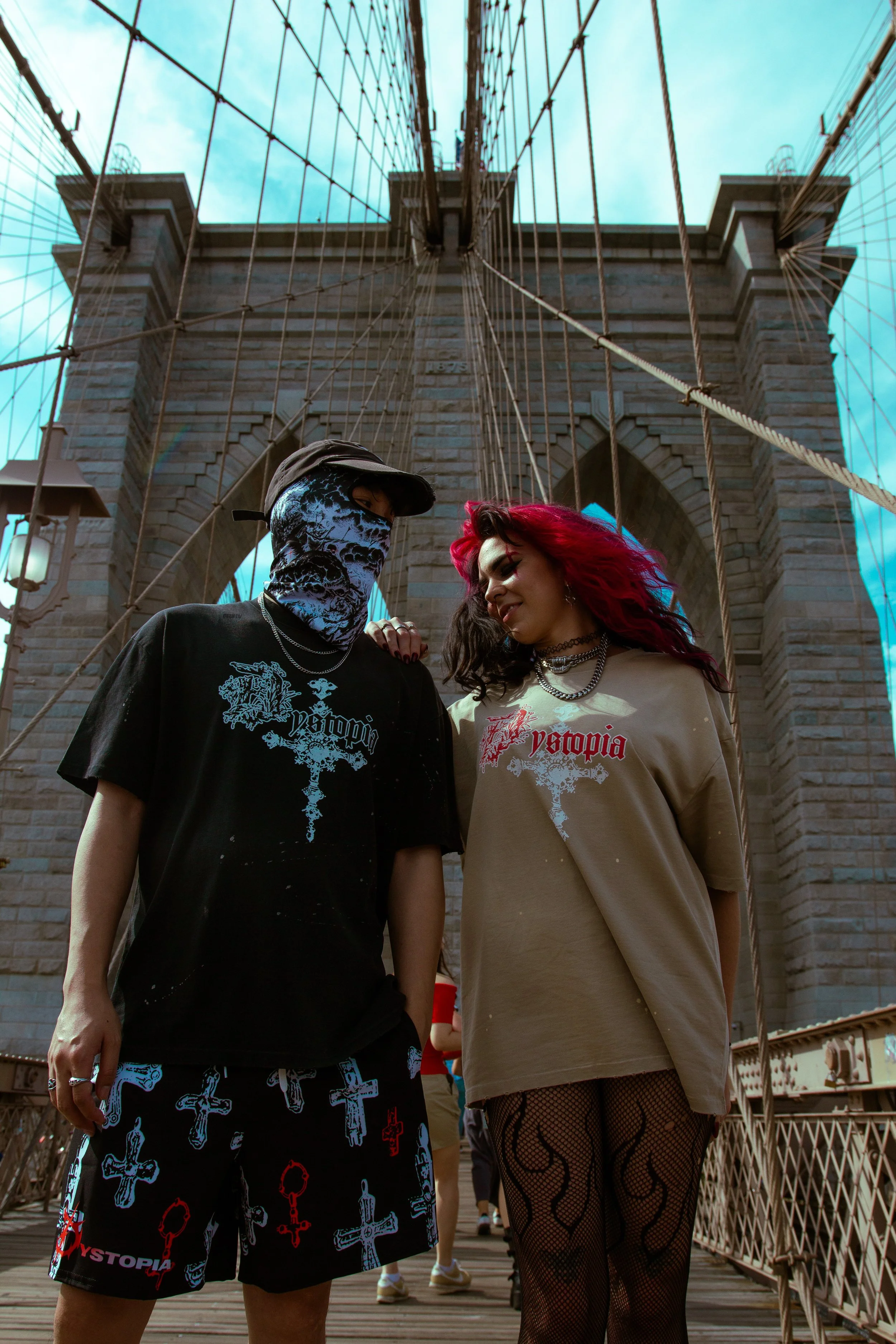 Two young people with alternative fashion standing on Brooklyn Bridge in New York City. The person on the left is wearing a face mask, black shirt, and shorts with cross and chain graphics. The person on the right has bright red hair, fishnet stockin