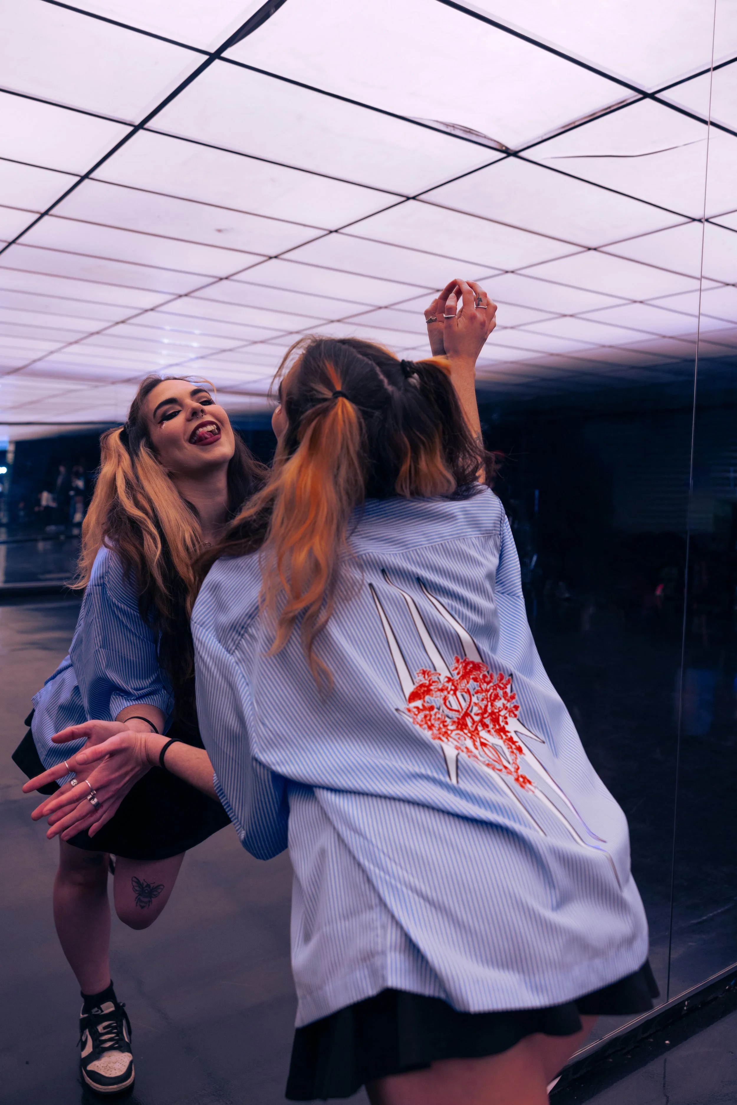Two young women with long hair and striped shirts having fun and taking selfies in front of a mirror in a modern, colorful indoor space.