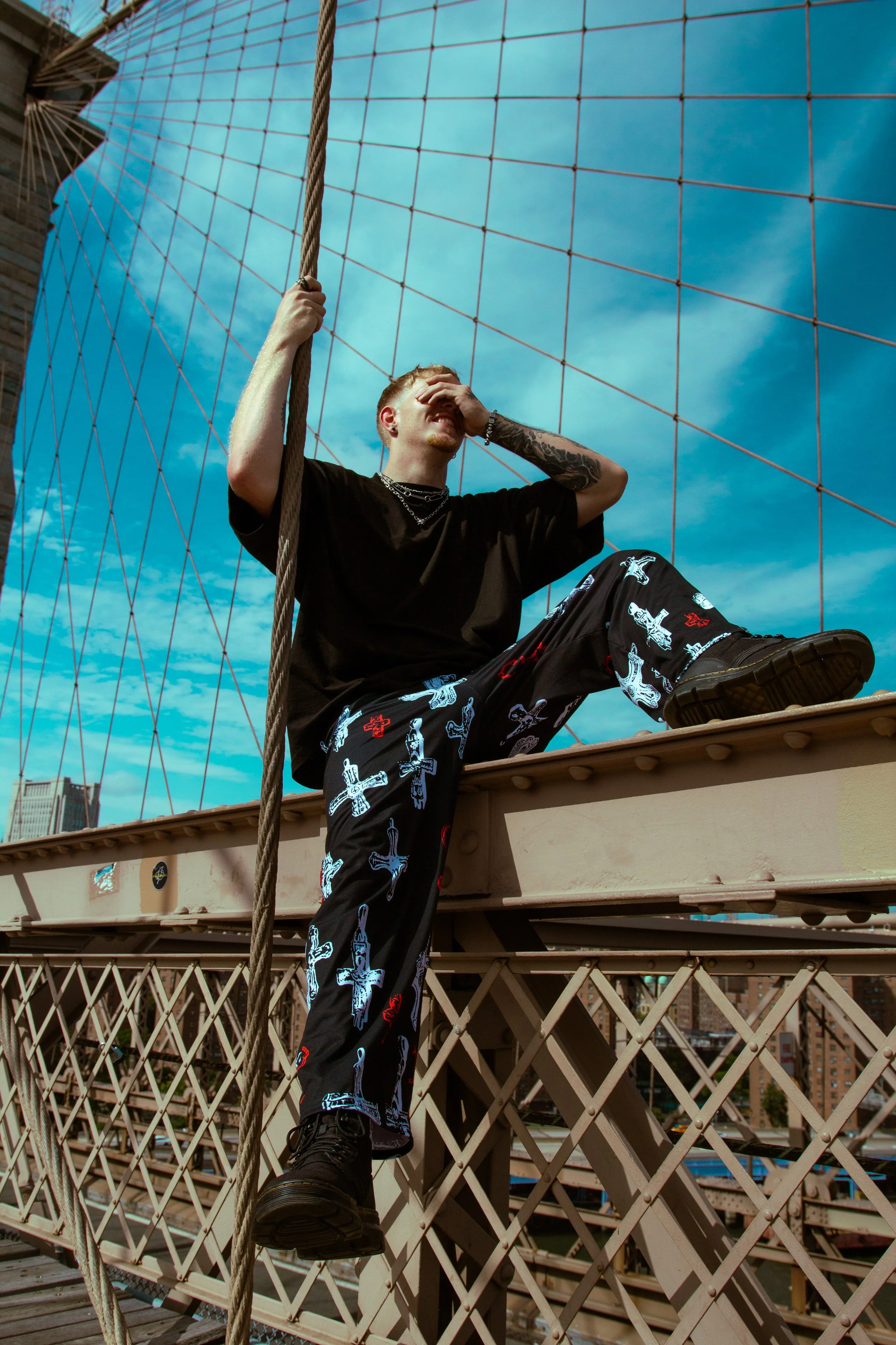 A young man sitting on a bridge in New York City, covering his eyes with one hand and holding a rope with the other, with skyscrapers and blue sky in the background.