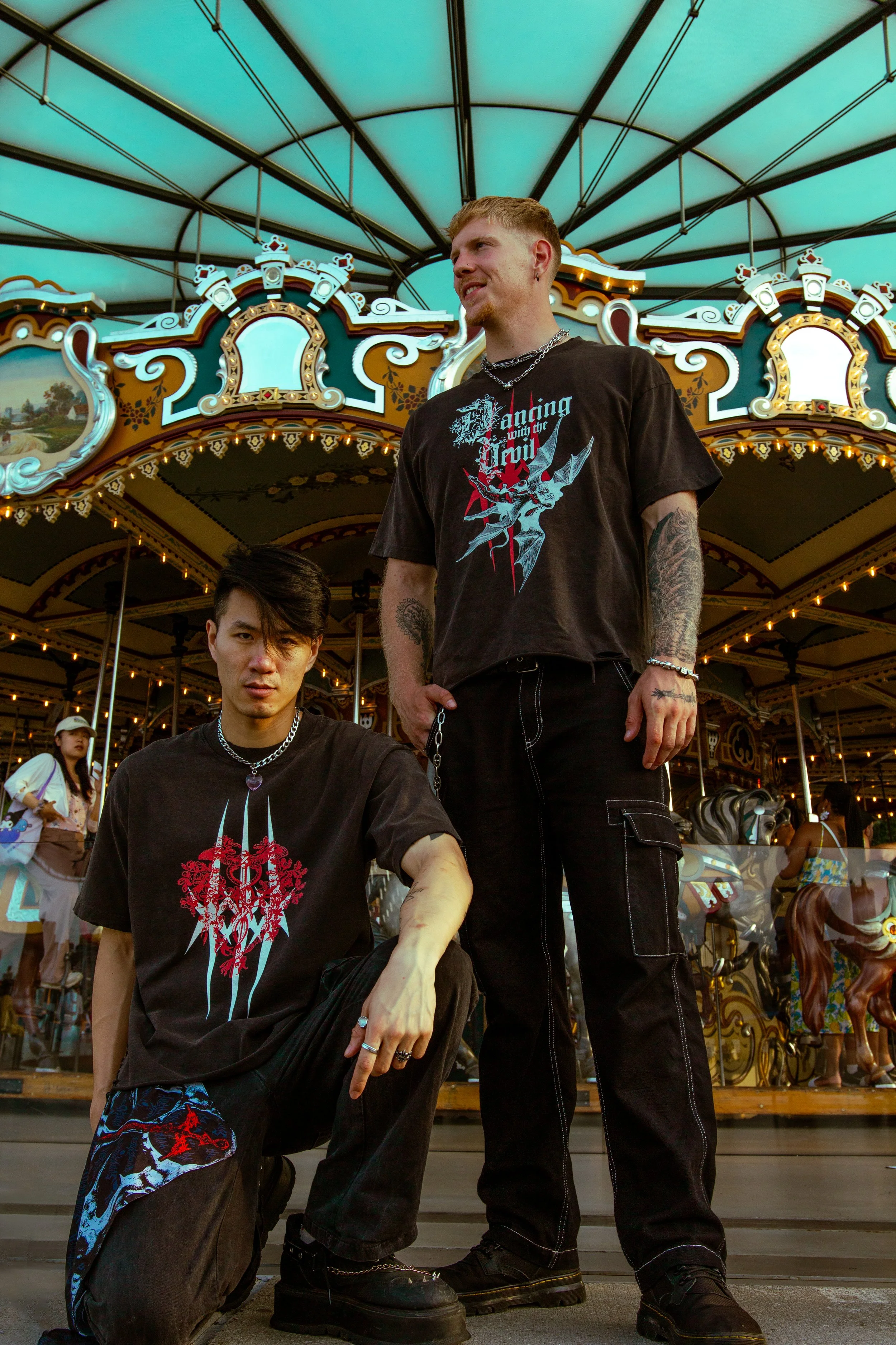 Two young men with tattoos and dark clothing posing in front of a carousel at an amusement park, with a canopy overhead and carousel horses in the background.