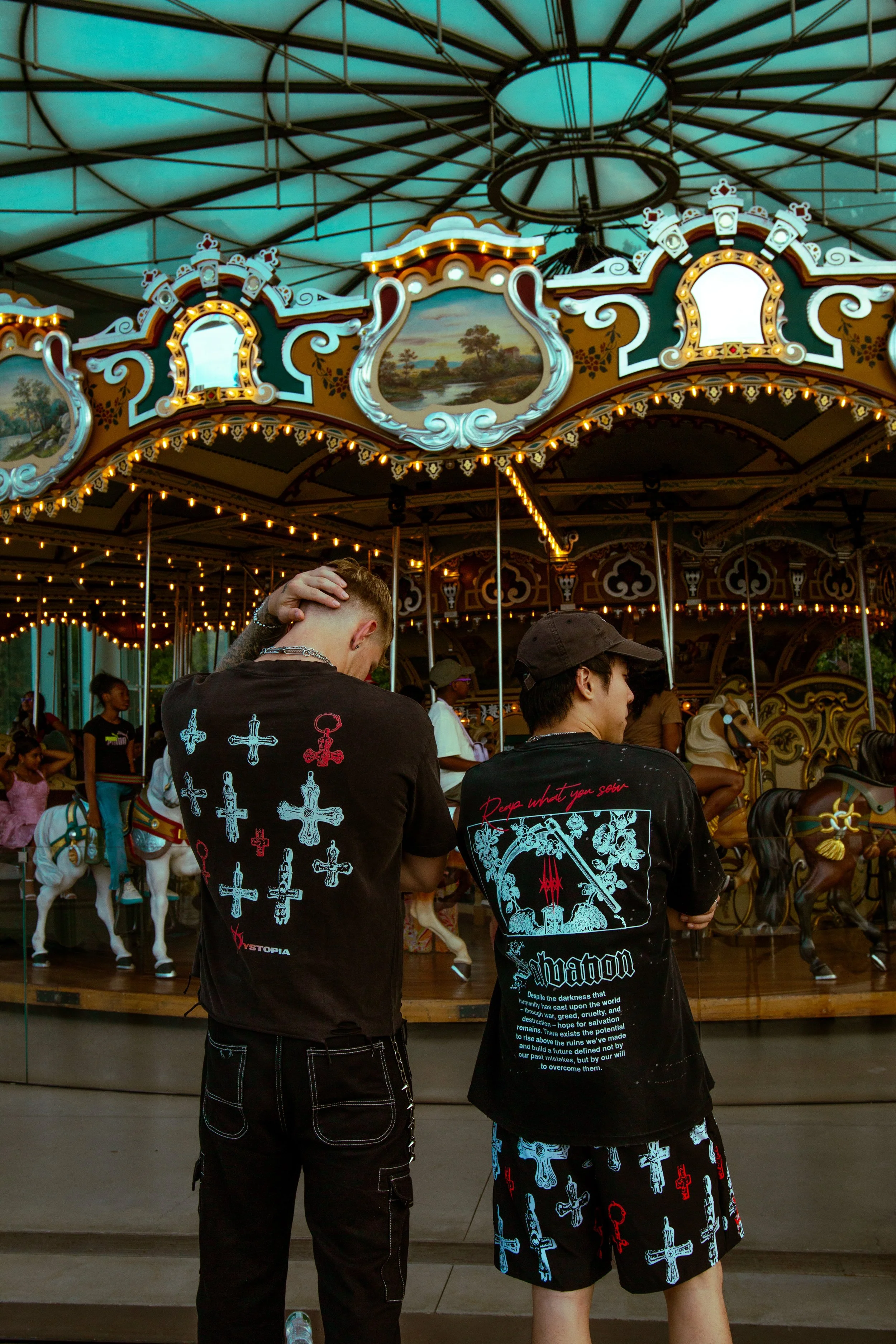 Two young men stand in front of a vintage carousel with ornate decorations and painted scenes, with children riding carousel horses in the background at an amusement park.