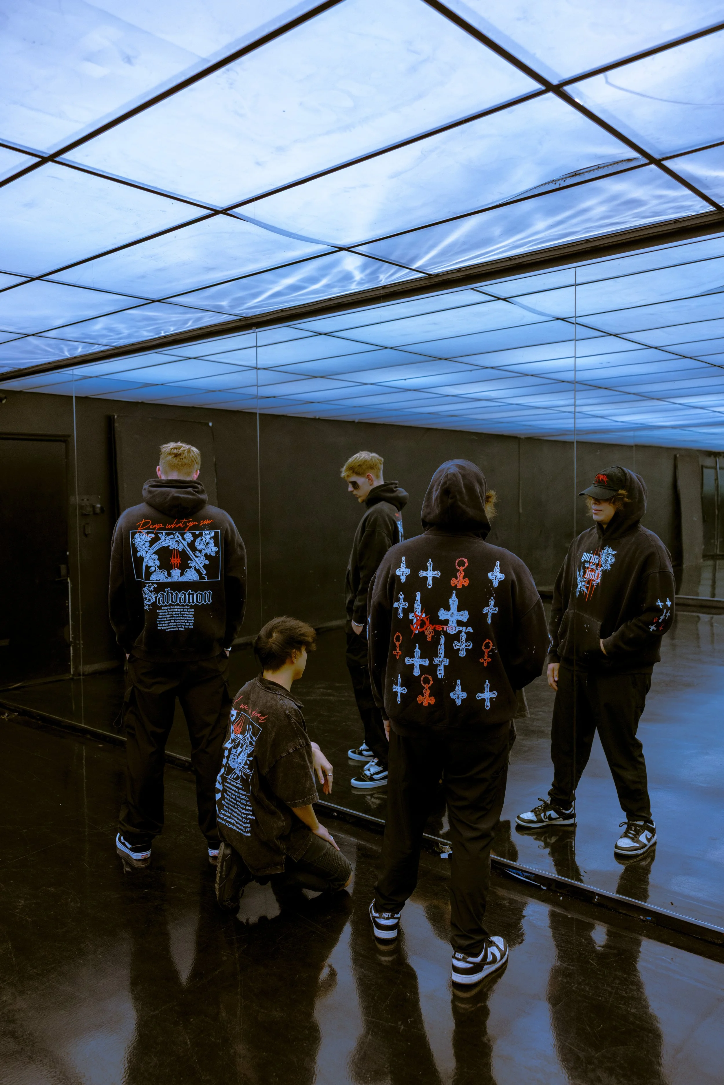 Group of young men dressed in black hoodies and sneakers standing in an indoor room with a reflective black floor and mirrored wall.